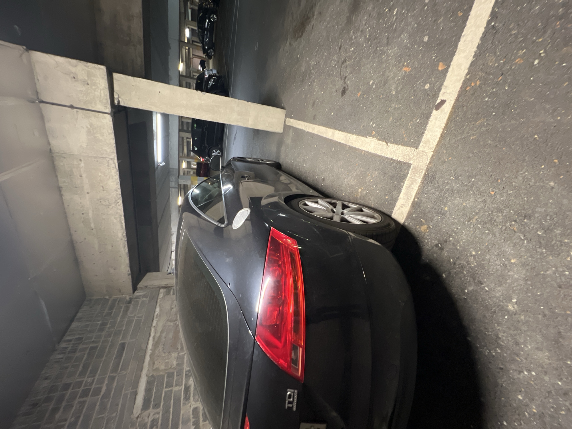The image shows a dark-colored Audi TT parked in an underground parking garage in an unknown city. The car is parked near a concrete support pillar and partially within parking space lines. The parking garage appears to be dimly lit, with several other vehicles parked further down the row. The car is positioned at an angle.