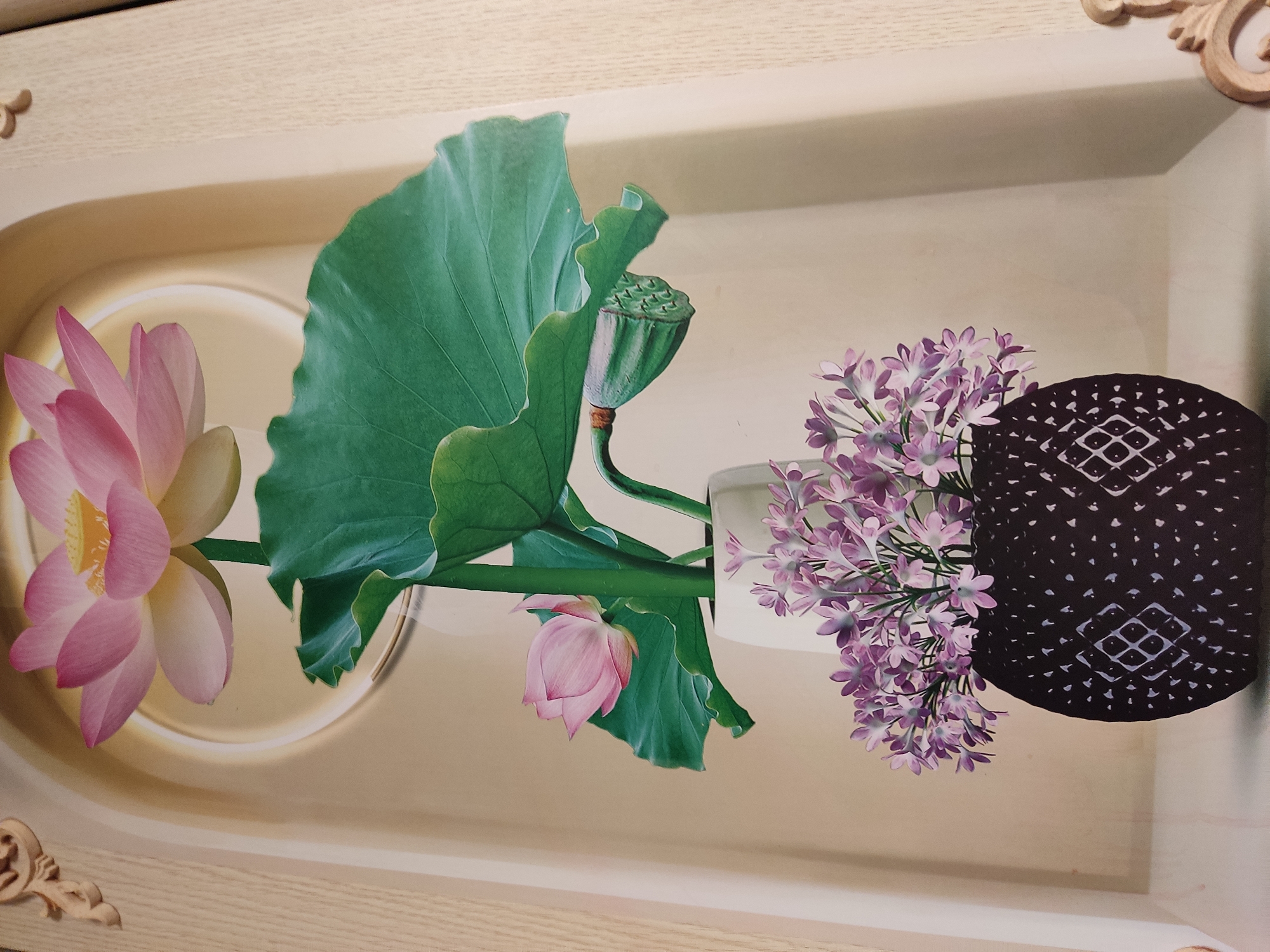 A vertical artwork depicts a still life of flowers in a vase against a neutral, recessed background. At the top, a large, fully bloomed pink lotus flower with yellow pistils is prominently displayed. Below it, a closed pink lotus bud is nestled among large, green lotus leaves. A seed pod with visible seeds hangs from a stem. These elements appear to be arranged in a tall, white vase. At the bottom of the composition, a dark, textured vase is filled with a bouquet of small, light purple flowers. The overall scene is framed by a decorative, light-colored border, possibly representing wood or a molding. There is no indication of the city of Noginsk, Russia, in the image.