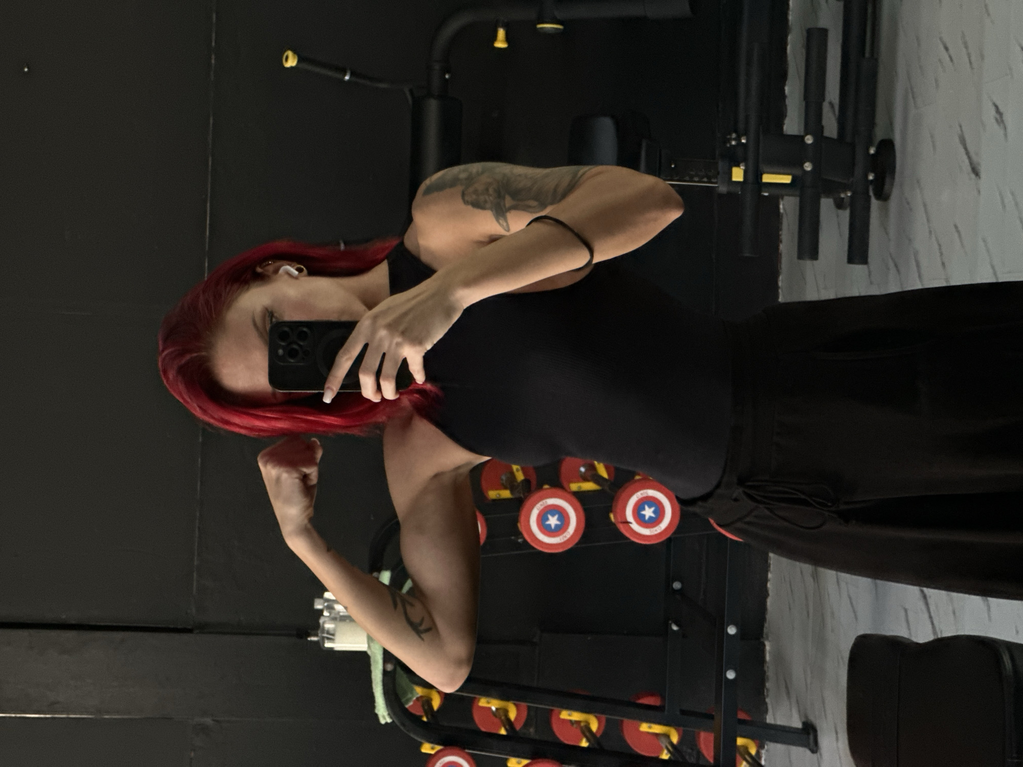 A woman with red hair is taking a selfie in a gym. She is wearing a black tank top and dark pants. The gym is located in Tibubeneng, Indonesia. Exercise equipment including weight racks with weights are visible behind her.