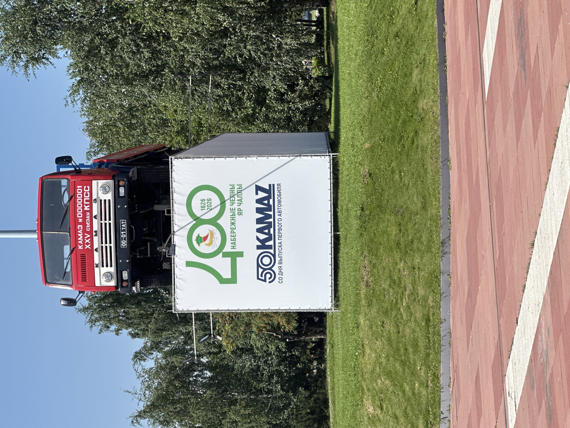 In the city of Naberezhnye Chelny, Russia, a red KAMAZ truck, identified by the license plate "00-01 TAT" and the inscription "КАМАЗ №0000001 XXV СЪЕЗДУ КПСС", is displayed atop a large white banner. The banner features a green "400" logo with a depiction of a boat and the text "1626 2026 НАБЕРЕЖНЫЕ ЧЕЛНЫ ЯР ЧАЛЛЫ". Below this, the KAMAZ logo is prominent with the text "50 KAMAZ СО ДНЯ ВЫПУСКА ПЕРВОГО АВТОМОБИЛЯ". The monument is situated on a grassy hill with trees in the background under a clear blue sky. The foreground shows a red and white tiled pathway. There are no people visible in the image.