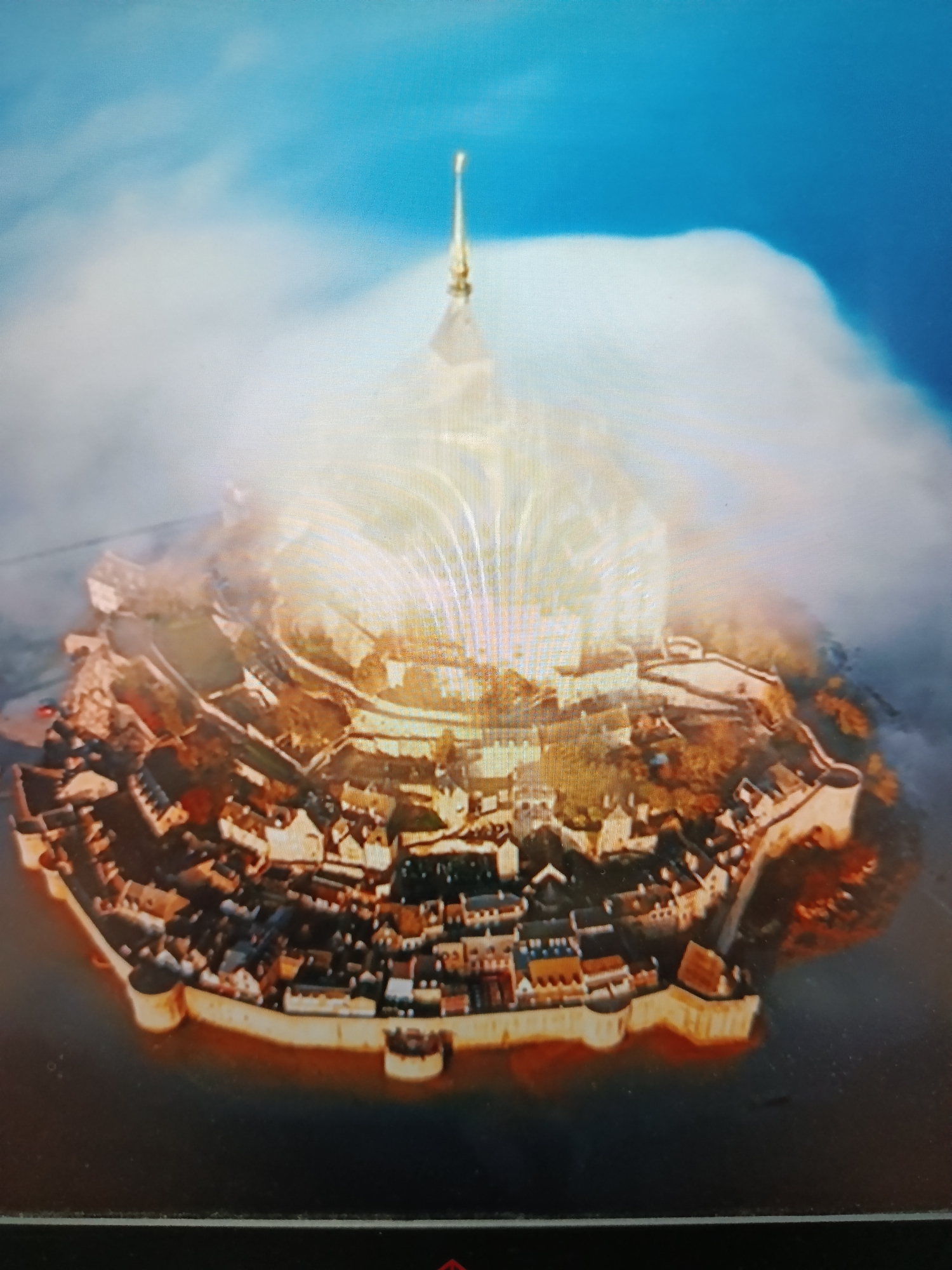 This media file displays an aerial view of a fortified island settlement, possibly Mont Saint-Michel in France, despite the provided location context of Tashkent, Uzbekistan. The image showcases a circular stone wall surrounding a densely packed village of buildings. Rising from the center of the island is a tall, spire-topped structure, likely a church or abbey, bathed in a bright, ethereal light. Wisps of clouds or mist encircle the island, adding a dramatic and almost mystical atmosphere. The surrounding water is dark and reflects the warm tones of the settlement. The scene is illuminated by what appears to be sunlight, possibly during sunrise or sunset, given the golden glow. There are no discernible people or text in the image. The overall impression is one of historical grandeur and isolation.