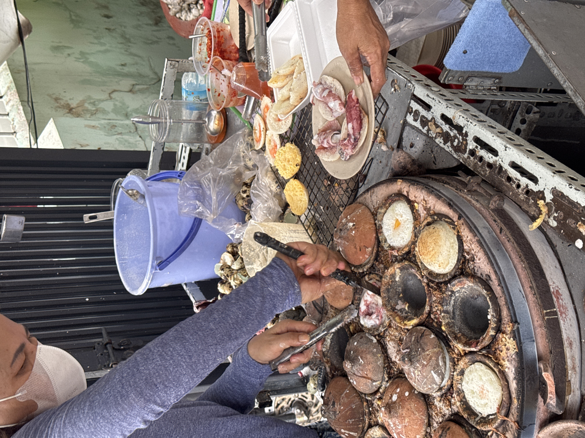 A street food vendor in Bac Nha Trang Ward, Vietnam, prepares bánh căn (mini savory pancakes) on a specialized cast-iron griddle with hemispherical molds. The vendor, wearing a face mask and a blue long-sleeved shirt, uses two metal tools to remove the cooked pancakes from the molds, which are filled with ingredients like eggs and meat. A person hands them cooked bánh căn with squid on top using a Styrofoam container and plate. A rack of toppings and condiments is visible, including containers of chili sauce, a blue bucket, and various pre-made pancakes. The cooking station is weathered and shows signs of frequent use.