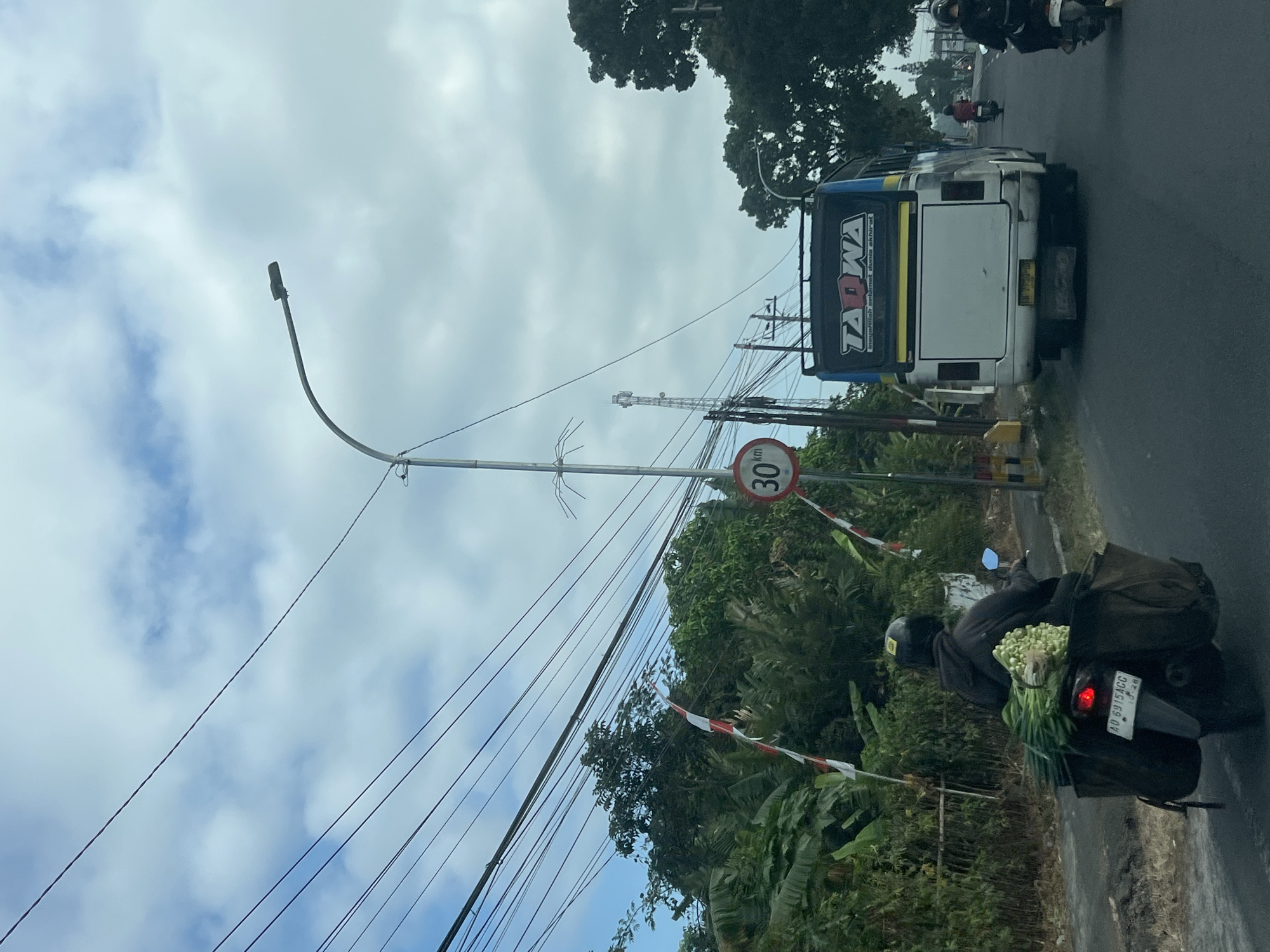 This is a medium-distance, eye-level shot of a road in Badran, Indonesia. The sky is overcast with scattered clouds. The road is paved and appears to be a two-lane road with traffic moving away from the viewer. On the left side of the road, there is a row of trees and vegetation, with electrical wires strung overhead. A person on a dark-colored motorcycle, wearing a dark hoodie and a helmet, is traveling in the foreground. The motorcycle is equipped with a large, dark brown bag attached to the rear, containing a bunch of green vegetables, including what appear to be leeks and onions. The motorcycle's license plate reads "AD 6915 ACC". To the right of the motorcycle, there is a white bus with blue and yellow accents, bearing the text "TADWA" on its rear. In front of the bus, a smaller red and white Indonesian flag is visible. Further down the road, two more motorcycles are visible, one red and one dark. A speed limit sign indicating "30 km/h" is mounted on a pole on the left side of the road.