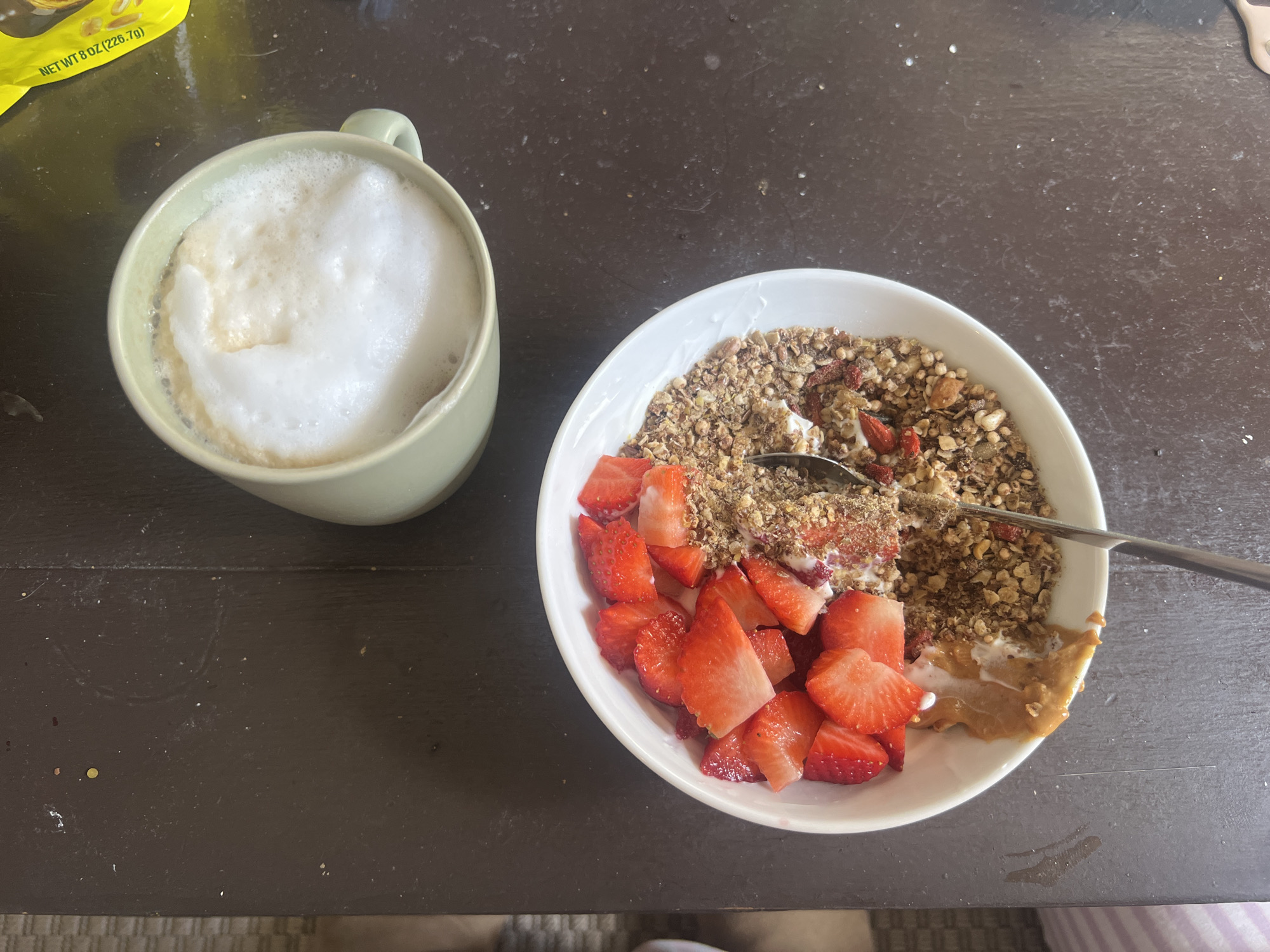 On a dark wooden surface are two food items. To the left is a light green mug filled with a foamy beverage. To the right is a white bowl filled with yogurt, granola, cut strawberries, and a spoonful of peanut butter. A metal spoon rests in the bowl. A small portion of a yellow package is visible in the upper left corner. The photograph appears to have been taken indoors. There is no evidence to suggest the location is in Paris, France.