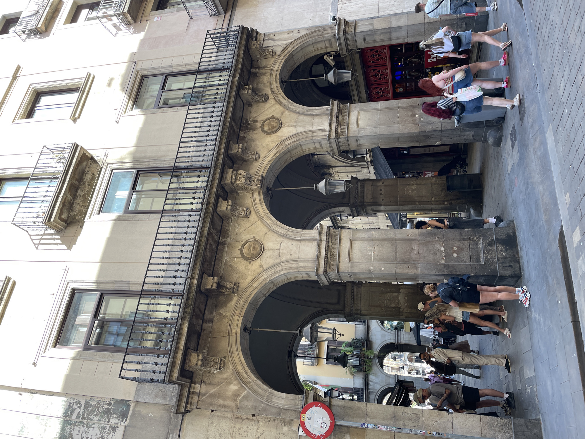 The media file captures a vibrant street scene in Barcelona, Spain, dominated by historic architecture under bright daylight. The central focus is a multi-story building featuring an elaborate stone facade with three prominent, ornate arches forming a covered walkway at street level. Above these arches, the building has several windows with dark frames and intricate wrought-iron balconies extending from the upper floors.
Numerous pedestrians populate the street below the arches and in the sunlit square in front of the building. People are seen walking, standing, and gathering, dressed in casual summer attire, indicating everyday street activity rather than a specific event. Some individuals are carrying bags.
Notable details include the strong, direct sunlight creating stark contrasts and shadows across the building and pavement, suggesting a clear, sunny day, likely late morning or early afternoon. Two antique-style lantern streetlights hang from the ceiling of the arched passage. One of the arches features a circular stone plaque with text, partially visible as "PASAGE DEL MARTE" (likely Pasaje del Martí). Near a distinctive red, ornate door to the right, the numbers "21" and the offer "2x1" are discernible, suggesting a commercial establishment. A red "no entry" traffic sign is visible in the lower left corner. The architectural style, with its stone arches and wrought-iron details, evokes a traditional European urban setting.