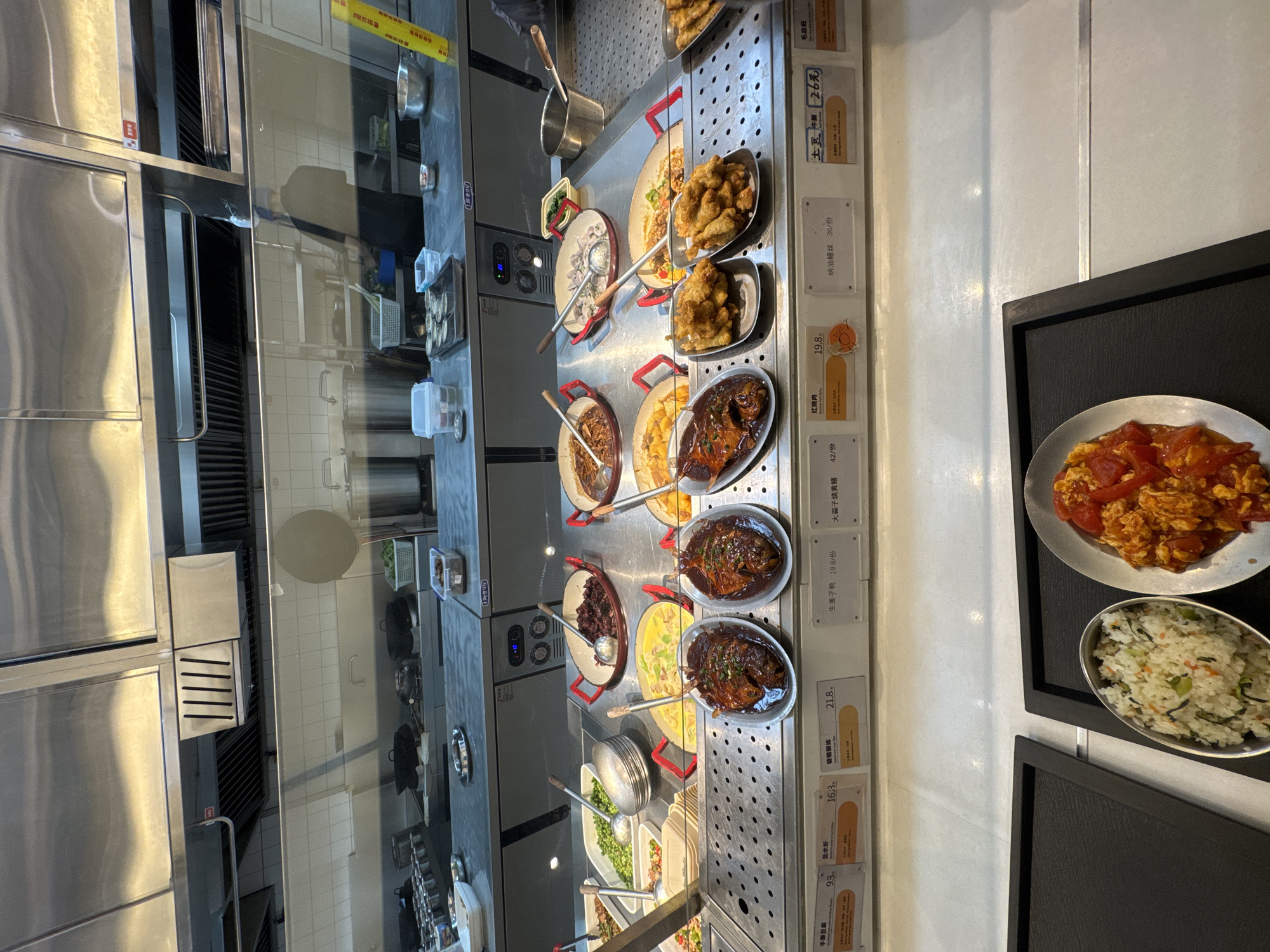 A food court is depicted in this image, likely in the Yangpu District of China, featuring a buffet-style selection of Chinese dishes. The foreground displays two black trays, one containing a silver oval dish of white rice mixed with green vegetables and another silver oval dish of scrambled eggs with tomatoes. Behind these trays, the main buffet line is visible. Various dishes are presented in metal and ceramic containers, including what appears to be braised pork, stir-fried edamame, and several saucy meat and vegetable preparations. Labels with Chinese characters and prices are affixed below each dish, indicating a self-service operation. In the background, stainless steel kitchen equipment, including exhaust hoods and cooking stations, is evident, suggesting an active food preparation area. The overall environment is clean and well-lit, typical of a modern cafeteria or food court setting.