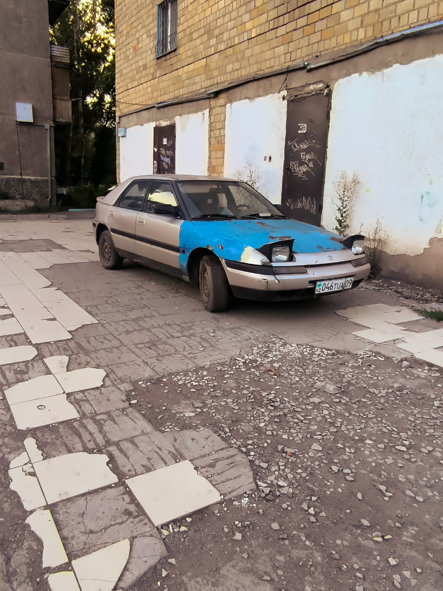 A silver and blue car with its headlights popped up is parked on a cracked, paved surface in front of a brick building. The car has a license plate that reads "KZ 046TUA 09". The building has large garage doors and a window with bars. There are trees and a smaller building to the left of the car. The ground is covered with debris and broken paving stones. The overall scene appears to be in an urban or industrial area.