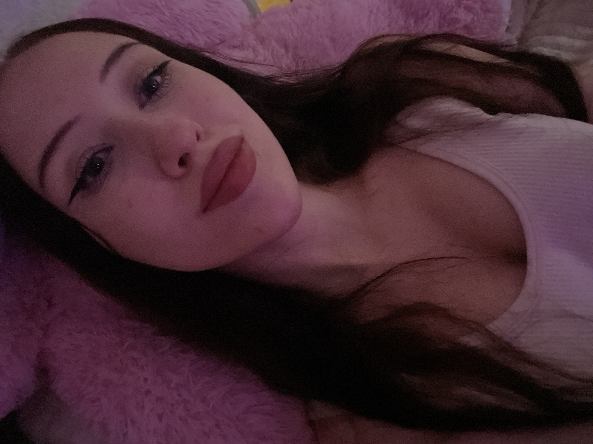 The image is a close-up, low-angle selfie of a young woman lying down, with her head tilted towards the camera. She has long, dark hair that drapes over her shoulder and a pink fluffy surface behind her. She is wearing a white ribbed tank top. Her makeup includes winged eyeliner and lip gloss. The lighting appears to be dim and pink-hued, possibly from an artificial source. There is no information in the image to suggest a location in Votkinsk, Russia.