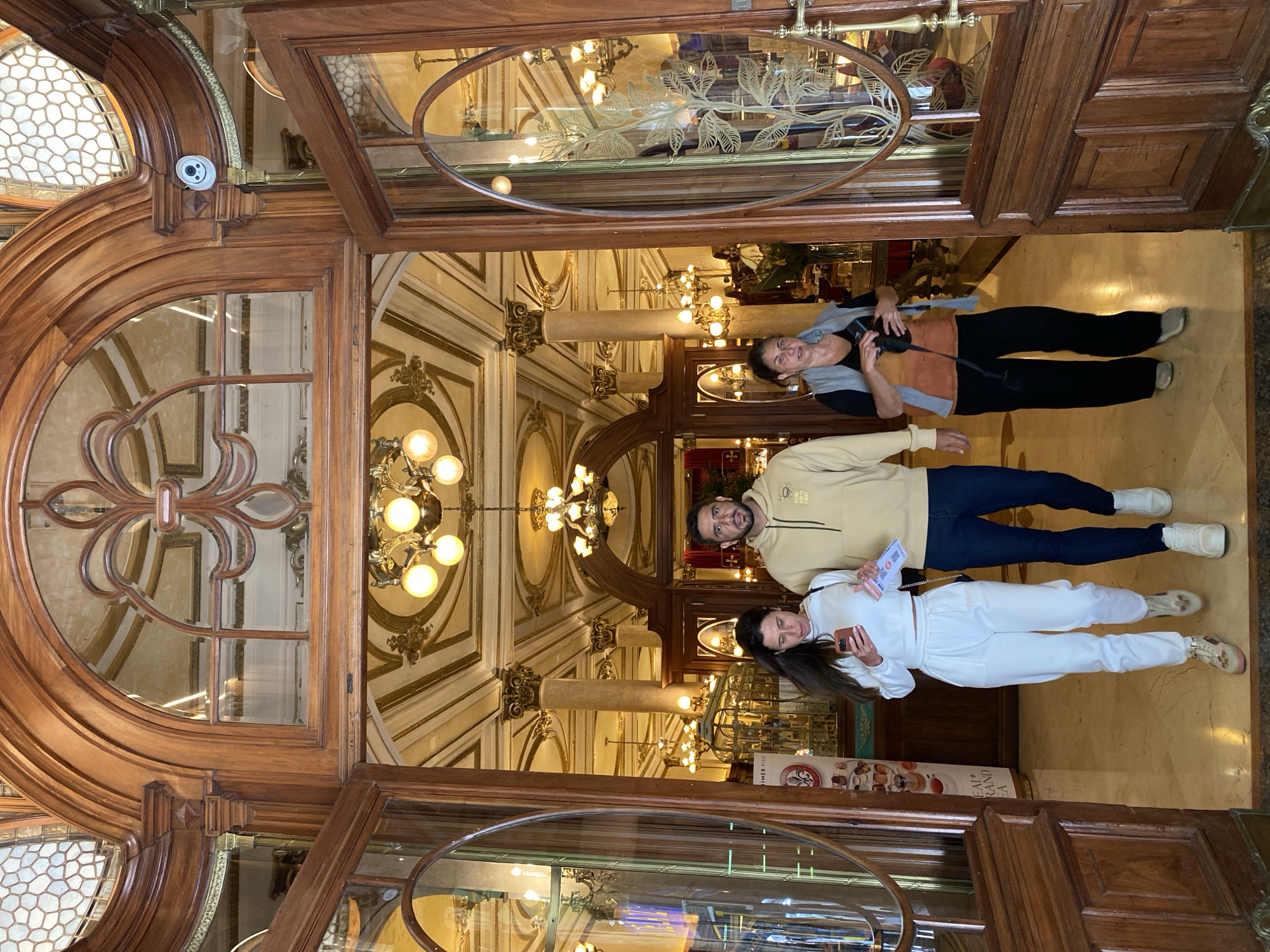 Three individuals, two women and one man, are captured posing for a photograph inside an opulent building, likely a grand cafe or historic establishment in Buenos Aires, Argentina. The image is taken from a slightly low angle, looking upwards towards the ornate entrance.
The man, positioned centrally, wears a light beige hoodie and dark pants, holding a small paper item. To his right, a woman in a white tracksuit and sneakers holds a smartphone. To his left, an older woman in black pants, a black top, and a grey vest holds what appears to be a camera or large phone. All three are looking directly at the camera with relaxed expressions.
The setting is characterized by elaborate wooden architectural details, including a richly carved doorway with arched and oval glass panels. Some of the glass features etched floral or leaf designs. High above, multiple chandeliers with exposed bulbs illuminate the space, casting a warm glow. The walls are adorned with decorative molding and gold accents, and tall pillars with ornate capitals are visible in the background. A small white security camera is discreetly mounted on a wooden arch in the upper left. The floor appears to be polished marble or stone.
Behind the woman in white, a display stand shows partially visible text, including "MER PISO" (likely "primer piso," meaning first floor) and "EAL+RAND EA," alongside illustrations of food and drinks, suggesting a menu or advertisement. The luxurious interior and period-style decor are strong indicators of a historic and upscale venue.