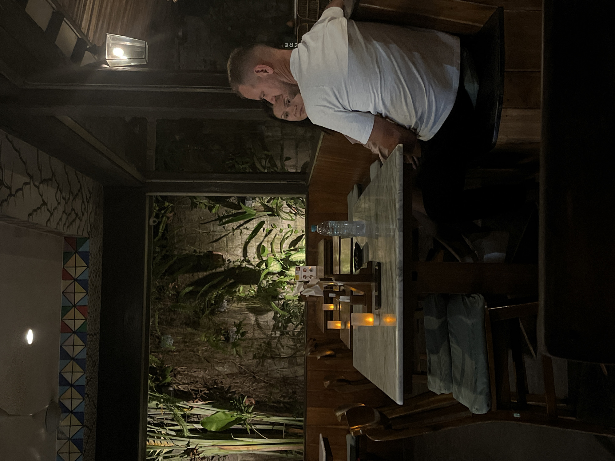 In the city of North Kuta, Indonesia, a dimly lit restaurant setting is depicted. A man with short blonde hair, wearing a white t-shirt and black pants, is seated at a table. He is looking away from the camera, and a woman with dark hair, wearing a pink top, is partially visible next to him. They are seated in a booth. In the foreground, a marble-topped table is visible with two small lit candles and a bottle of water. Behind the table is a window showing a lush, green wall of plants. The restaurant's decor includes a tiled border along the top of the wall and a lantern light fixture hanging from the ceiling. Wooden chairs with blue cushions are also visible around the tables. The atmosphere appears to be serene and intimate.