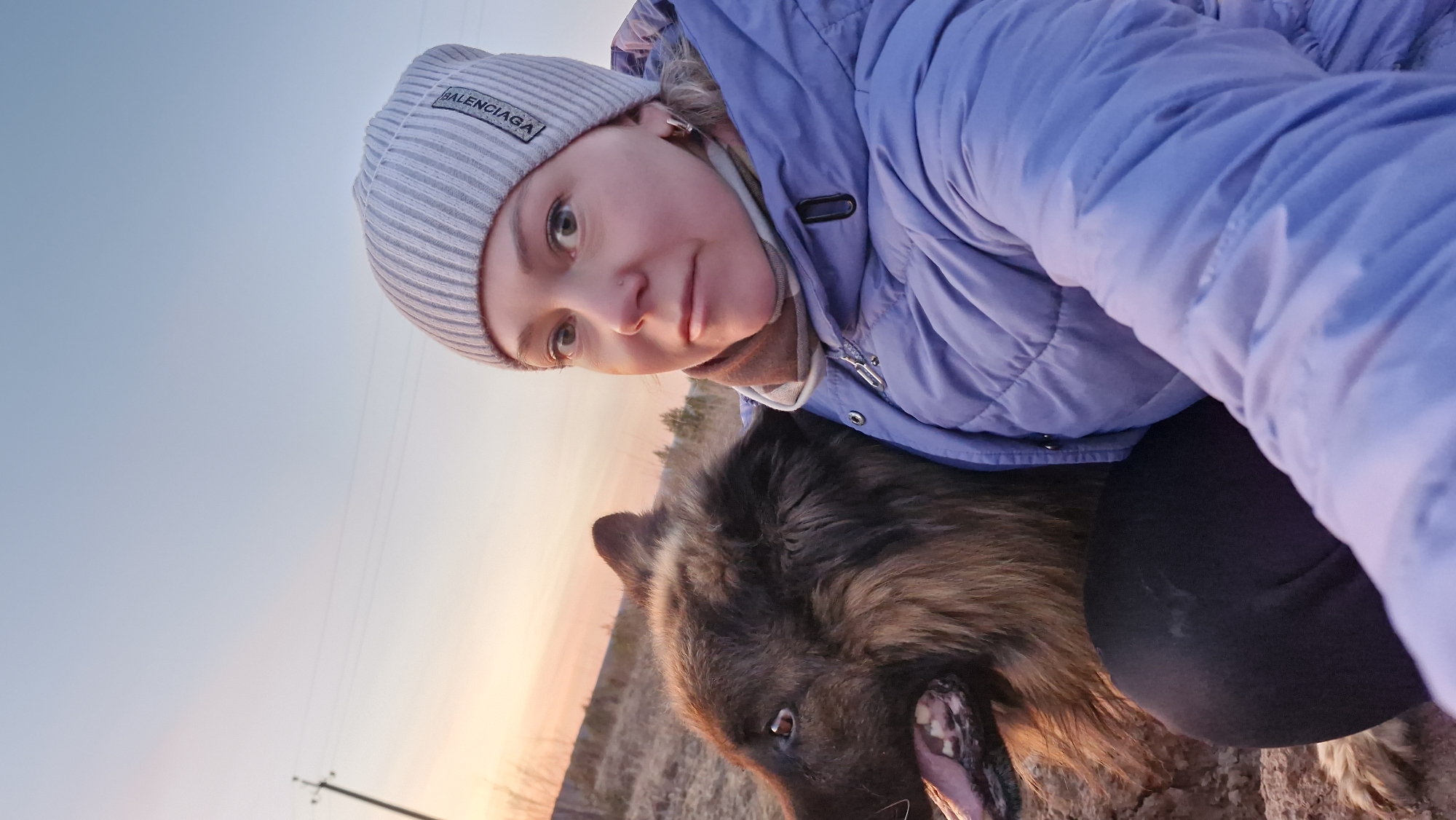 This tilted selfie, taken in Otchor, Russia, captures a woman and a large dog outdoors during either sunrise or sunset. The sky displays a soft gradient of orange and pink hues near the horizon, transitioning to a pale blue overhead.
The woman, positioned in the upper right, looks directly at the camera with a neutral to slightly smiling expression. She is wearing a light purple puffer jacket and a light grey, striped beanie that features a black label with the text "BALENCIAGA." A checkered scarf in brown and beige tones is visible around her neck, suggesting cool weather.
Beside her, a large, shaggy-haired dog, possibly a German Shepherd or similar breed, is partly visible. The dog has dark brown and tan fur, and its mouth is open, revealing its tongue and teeth. Its head is close to the woman, suggesting companionship.
The background shows a sparse, outdoor environment with dry, light-colored grass or dirt. Distant, blurry trees or bushes can be seen near the horizon. A utility pole with power lines is visible in the lower left portion of the frame. The intimate, close-up shot focuses on the subjects against the colorful backdrop of the sky.