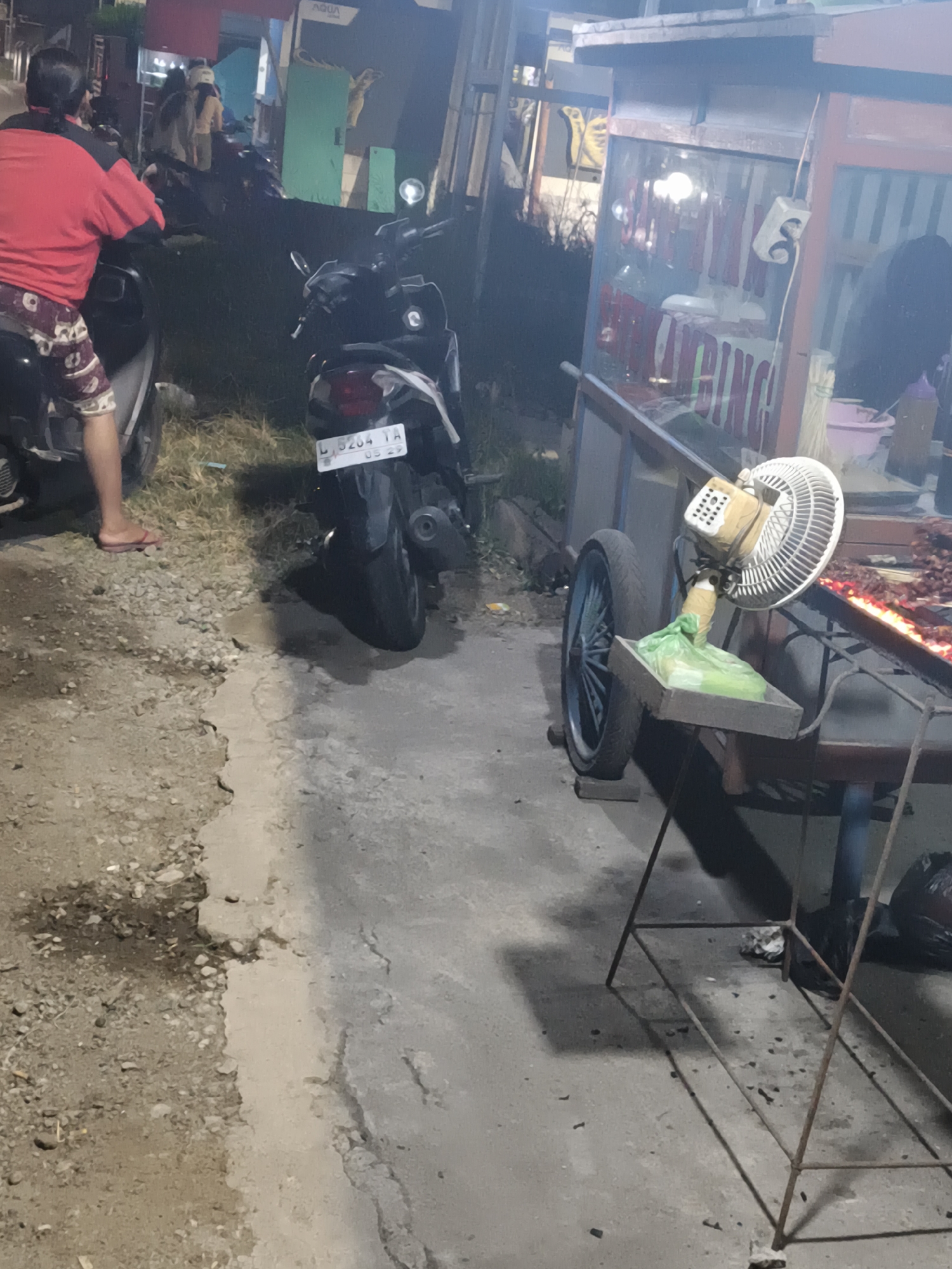 The image captures a dimly lit street scene at night, likely in Sidorejo, Indonesia, focusing on a food stall and its surroundings. In the foreground, a person wearing a red shirt and patterned shorts is seated on a motorcycle, viewed from behind. To the right, a mobile food cart is visible, displaying a grill with food cooking over it. A fan is attached to the cart, positioned to cool the area. Behind the food cart, a few people are seen standing. The environment appears to be an outdoor urban setting, with a paved surface and some vegetation. The lighting is predominantly artificial, coming from the stall and streetlights.
