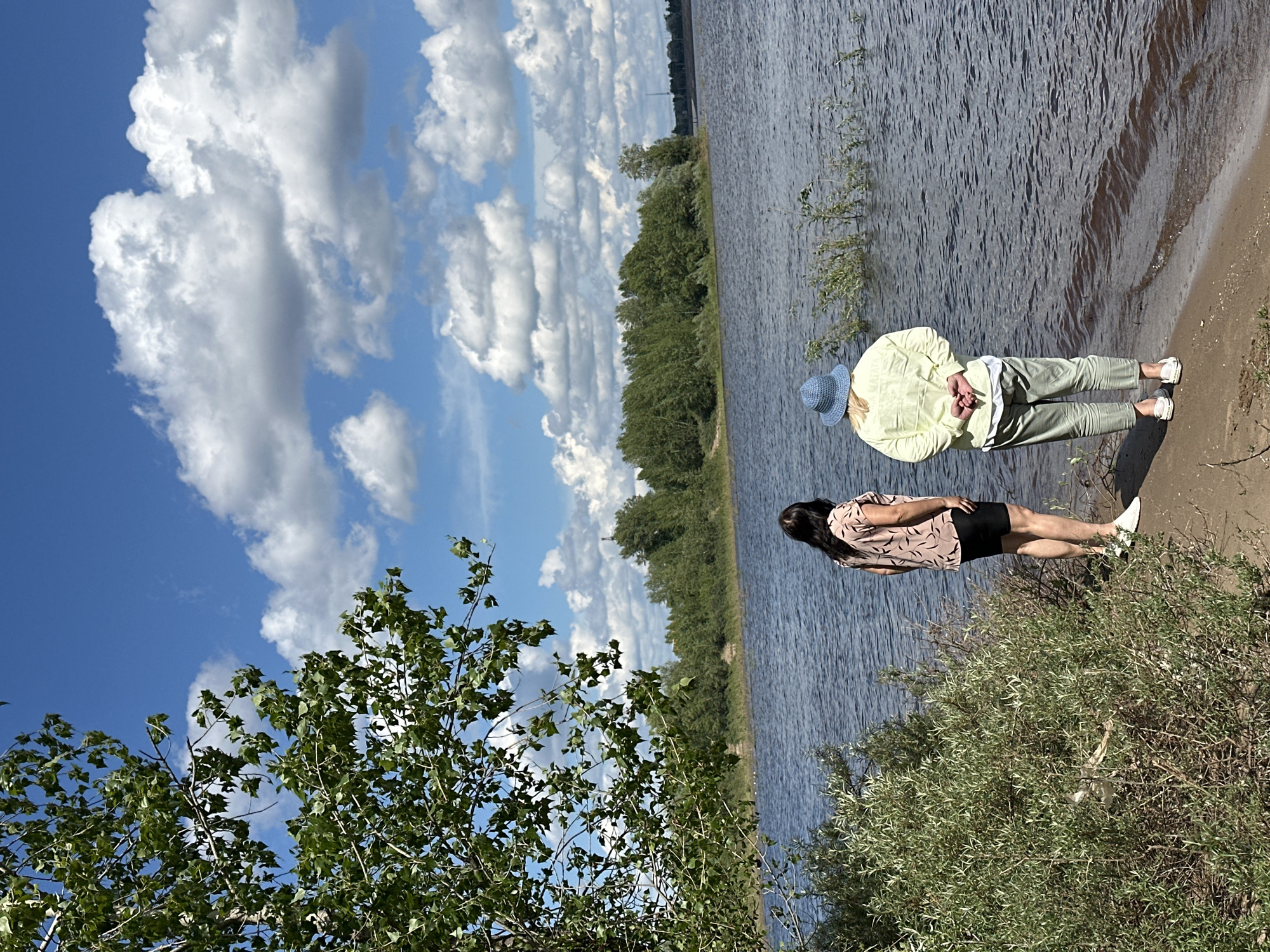 This image depicts a scenic view of the Kama River in Chaikovsky, Russia. Two women are standing on a sandy embankment, facing the water. The woman on the left has long, dark hair and is wearing a patterned short-sleeved shirt and black shorts. The woman on the right is wearing a light-colored long-sleeved jacket, light green pants, and a blue hat. She has her hands clasped behind her back. Lush green foliage frames the foreground, with the wide, calm expanse of the Kama River stretching out to the horizon. A dense line of trees borders the opposite bank of the river, and a bright blue sky with fluffy white clouds dominates the upper portion of the frame. The overall impression is one of peaceful contemplation by the water.