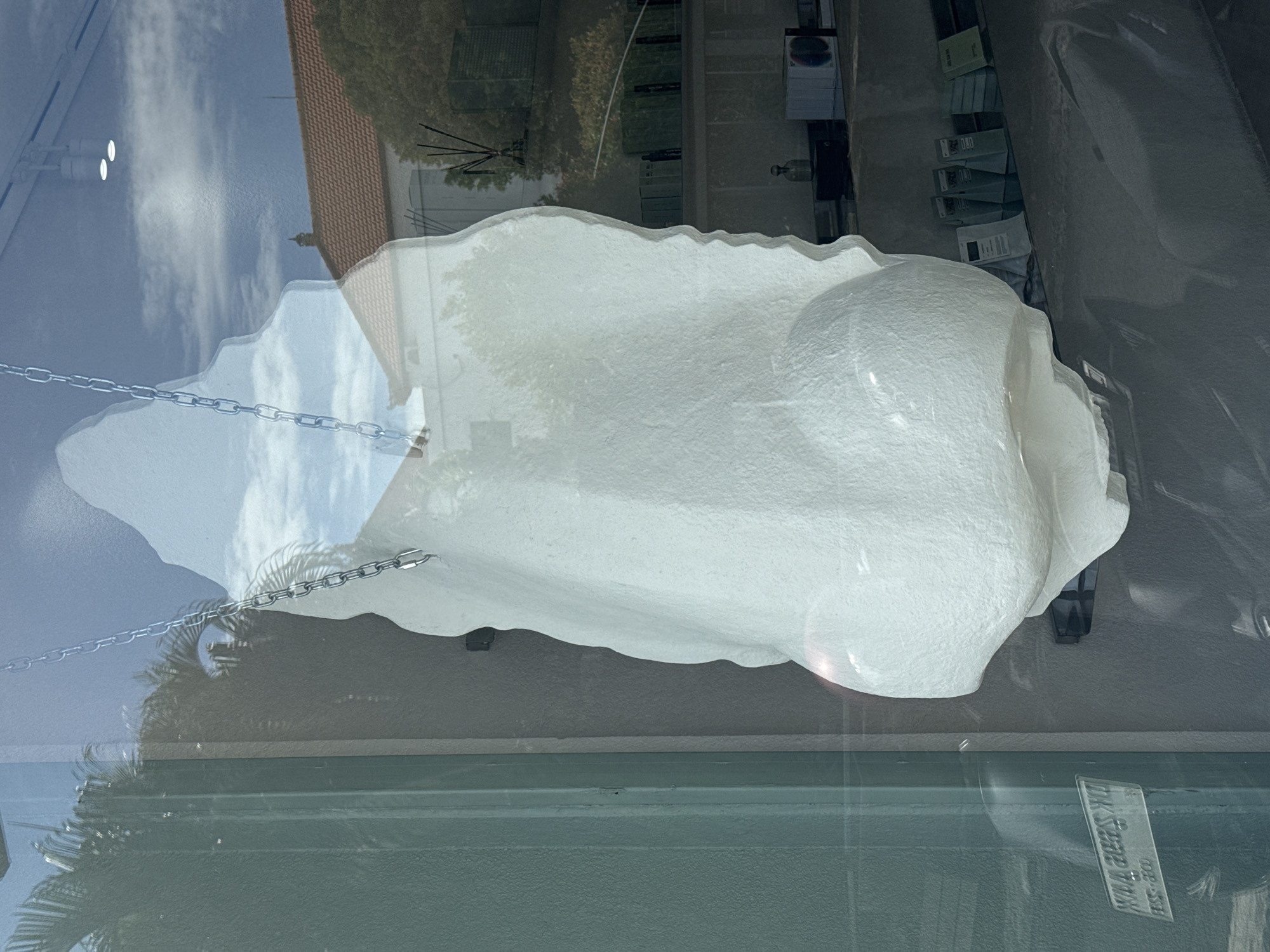 The image shows a white sculpture suspended by chains, as viewed through a glass window. Reflections in the window include buildings, trees, the sky, and possibly street lights. The sculpture is located in Unknown, Unknown.