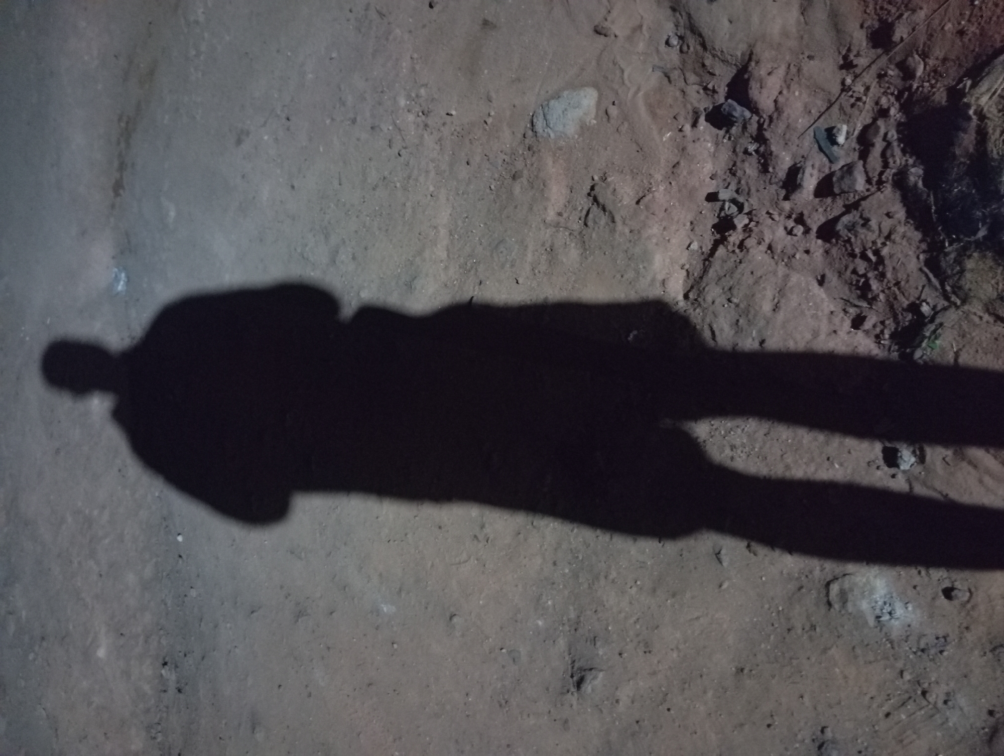 A shadow of a person is cast on a dirt and stone surface. The location, Nkpor, Nigeria, is an assumption based on limited information. No people or other subjects are directly visible, only the shadow. The environment consists of dirt, stones, and possibly gravel. No actions or interactions are observable.