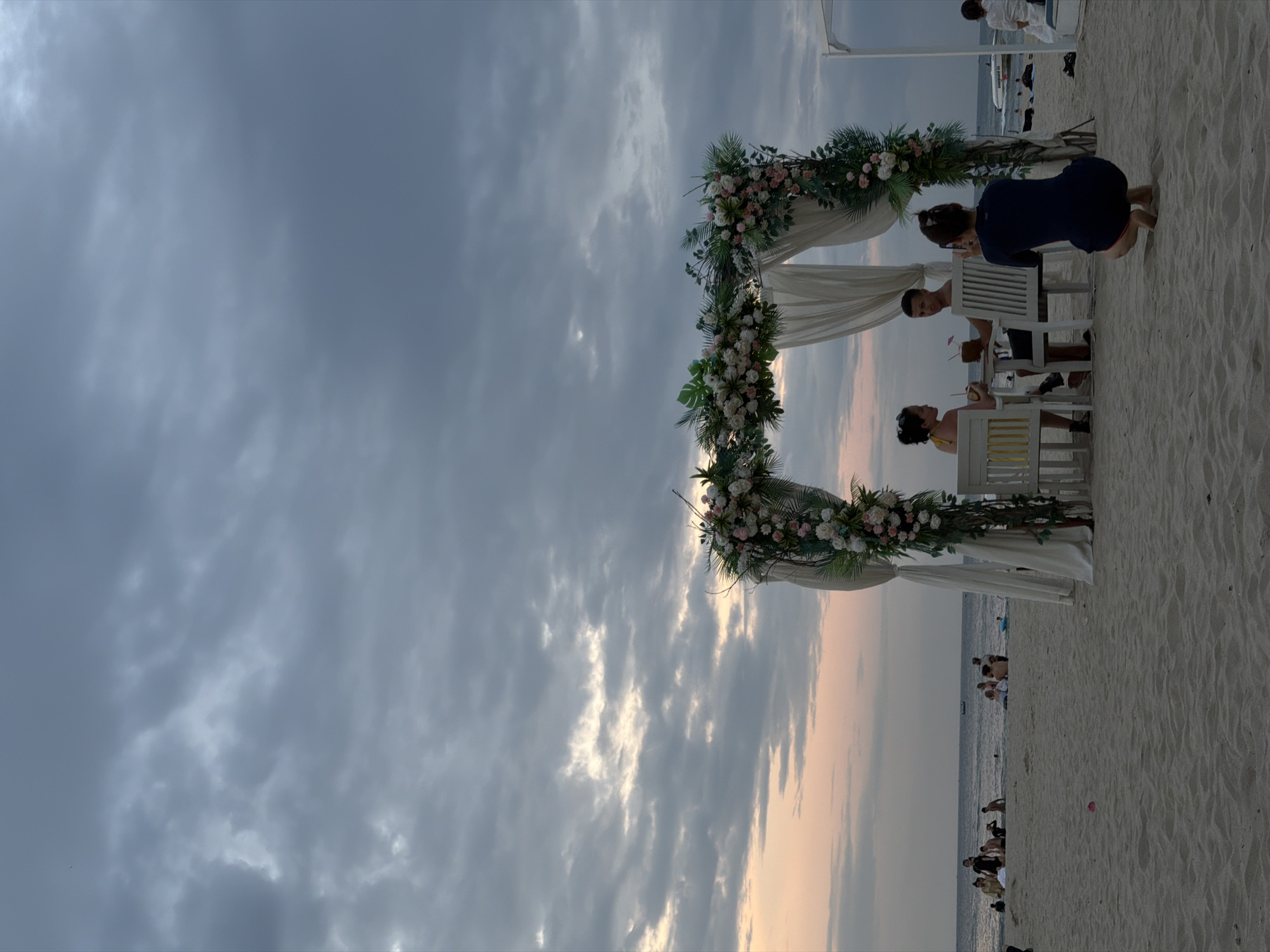 At dusk on a sandy beach in Đà Nẵng, Vietnam, a couple is seated at a table set up under a floral archway. The arch is decorated with an abundance of white and pink roses and green foliage. She, wearing a yellow top, sits on the left, facing him. He, in a dark t-shirt, is on the right, looking toward the camera. Both are holding drinks. To the right of the couple, a woman in a dark blue outfit is crouching and taking a photo of them with her phone. In the background, the ocean stretches to the horizon under a cloudy, pastel-hued sky. The beach is populated with several other individuals and groups enjoying the evening. A white canopy structure is visible to the far right.