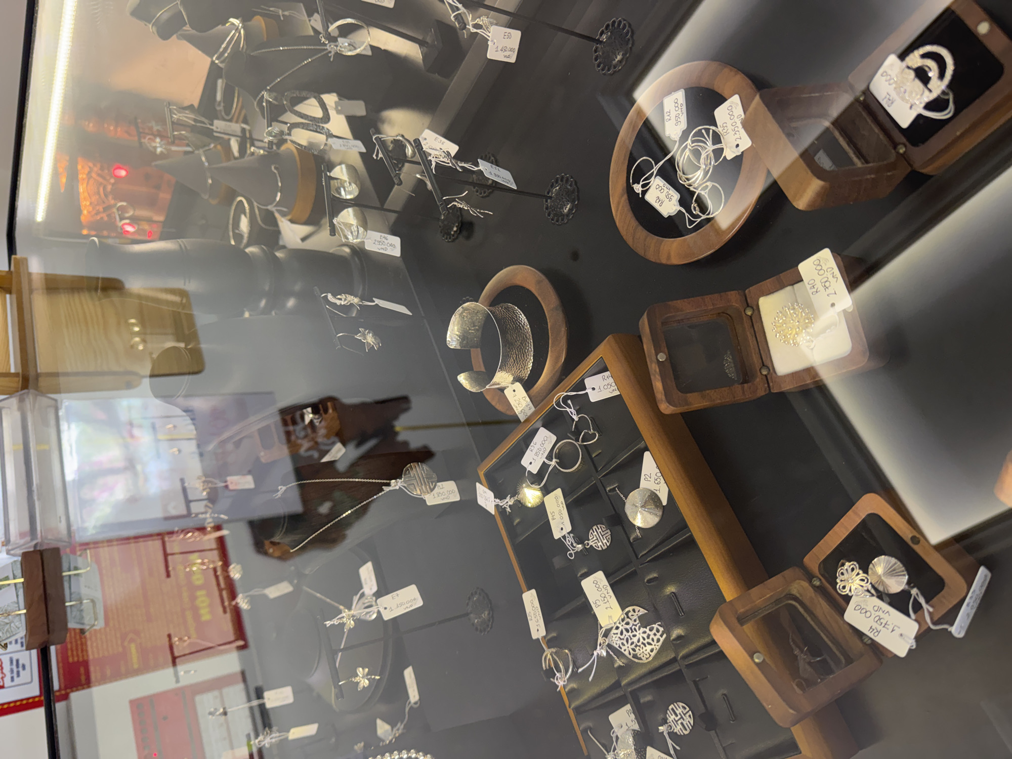 The image shows a jewelry display case, likely inside a store, with a variety of silver jewelry items on display, many with attached price tags in Vietnamese Dong (VND). The tags show amounts like "1.750.000 VND", "2.750.000 VND," suggesting this is located in Vietnam, specifically, based on the signs in the background, the location is potentially in the city of Đà Nẵng, Vietnam. There are rings, bracelets, necklaces, and earrings, arranged on black display surfaces. Some items are presented in small wooden boxes. The lighting is bright, reflecting off the glass of the display case.