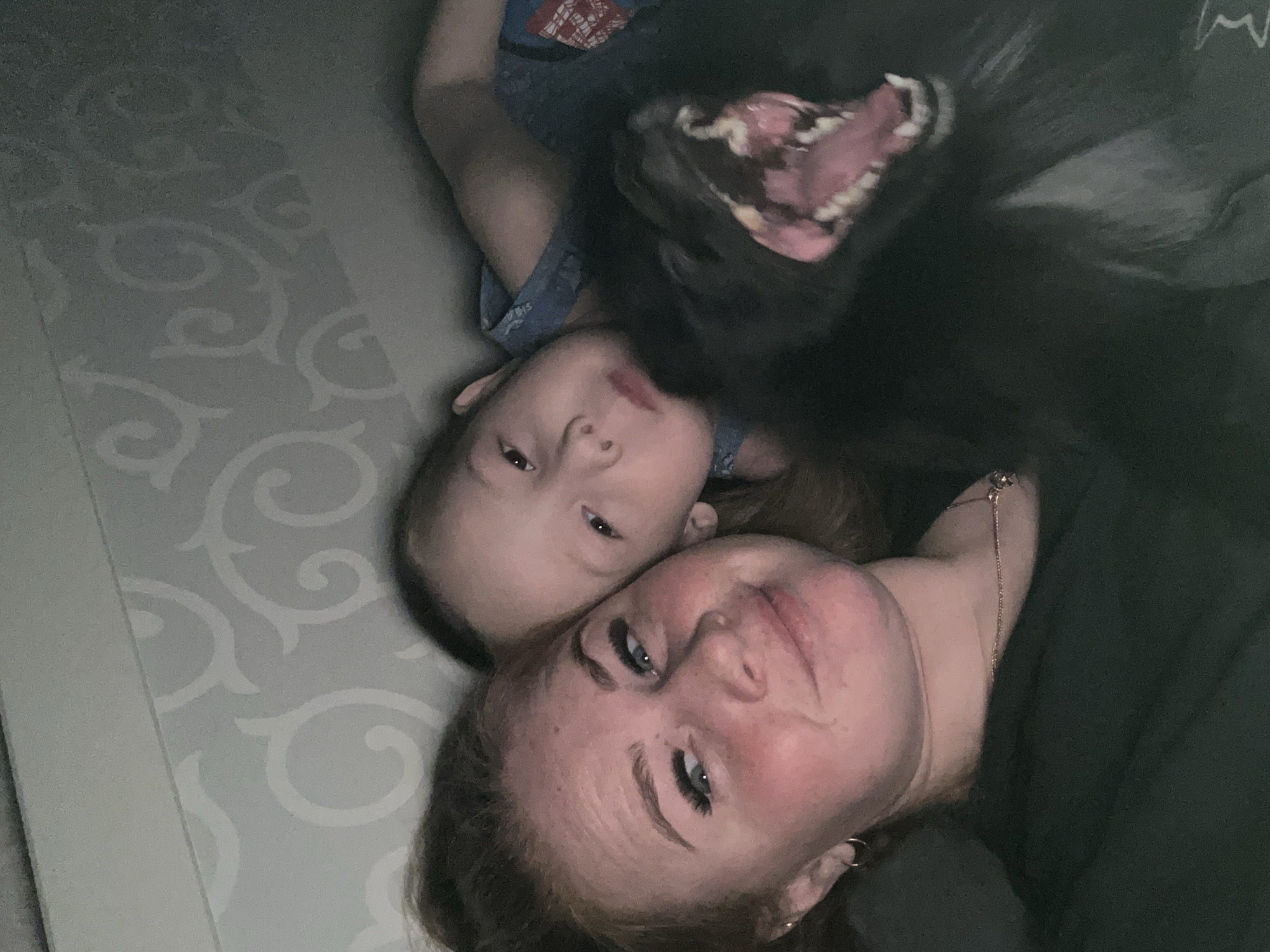 A woman, a child, and a black dog are in the frame. They are lying down indoors on a surface with a light-colored patterned covering. The woman is at the bottom of the frame, looking up. The child is above and to the left of the woman. The black dog is above and to the right of the child, appearing to open its mouth wide. No definitive evidence places the location in Chaikovsky, Russia.