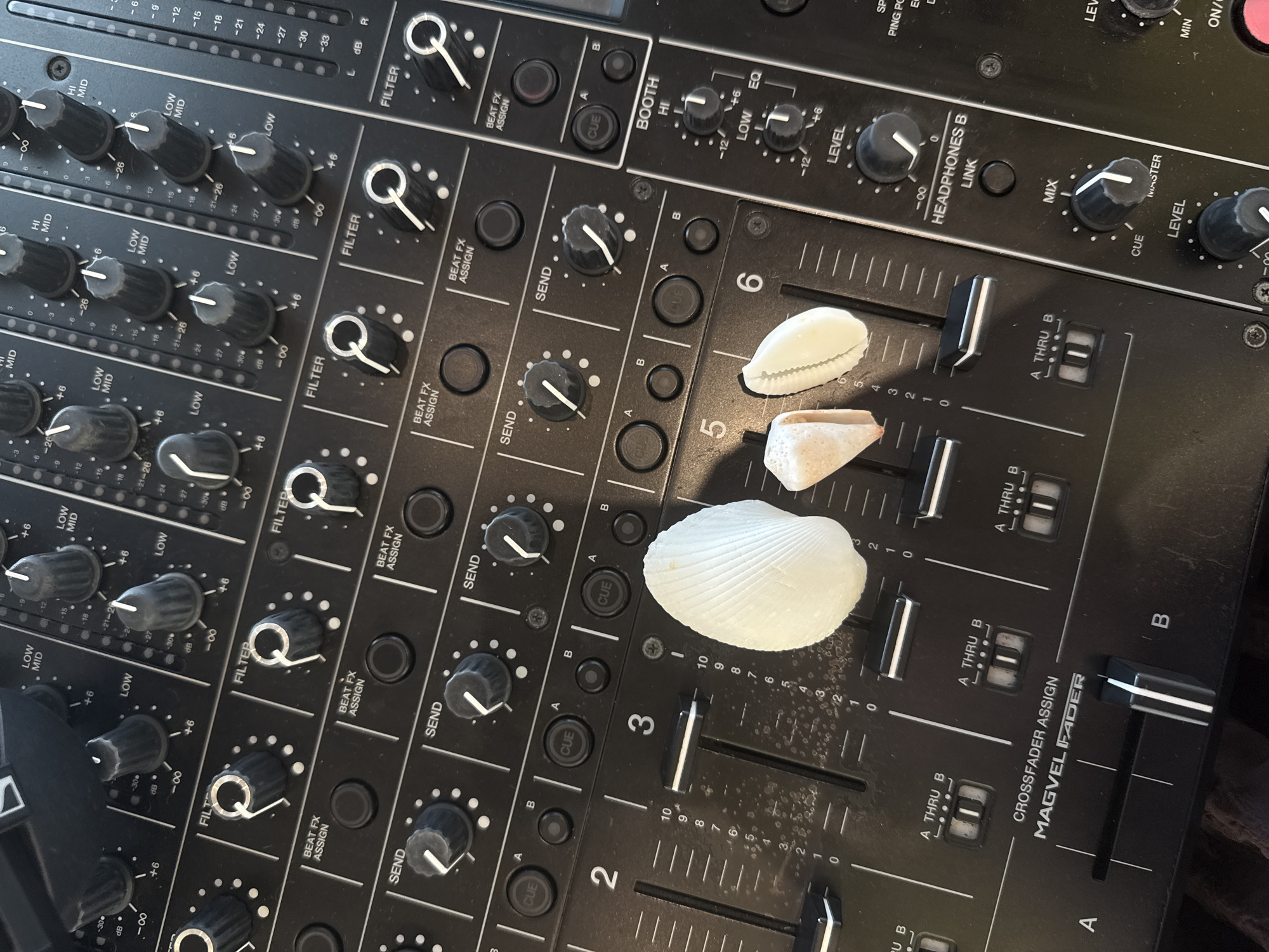The image shows a close-up, overhead view of a black DJ mixer. Three seashells are placed on the mixer's surface: a large, white, ribbed shell on the right, and two smaller, tan-colored shells to its left. The mixer has numerous knobs, sliders, and buttons, with labels such as "FILTER," "BEAT FX ASSIGN," "SEND," "CUE," "LEVEL," and "MIX." Faders are marked with numbers from 0 to 10. The environment is dark, with the focus on the detailed texture and labels of the DJ equipment. No people are visible. The location is noted as Unknown, Maldives.