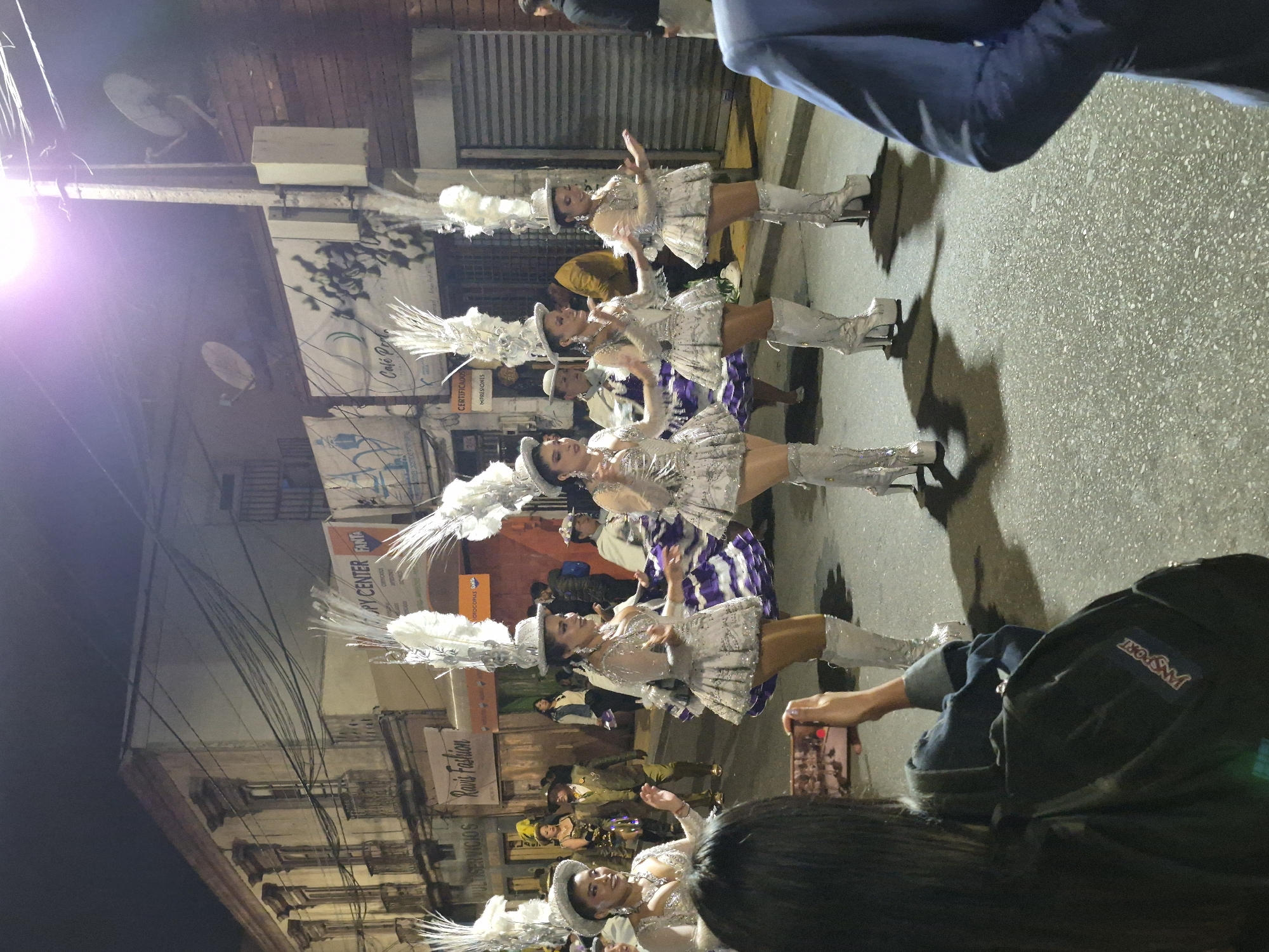 This is a nighttime, street-level, medium shot of a parade in Arica, Chile. The parade features several women in elaborate, sequined costumes with large white feather headdresses and tall, silver platform boots. They are all facing the viewer and appear to be dancing or posing. In the foreground, a person with dark hair is holding up a smartphone to record the parade. The street is paved, and there are buildings lining both sides of the street, some with businesses visible like "Ravi's Fashion" and "Café Perla". The overall atmosphere is festive and celebratory.