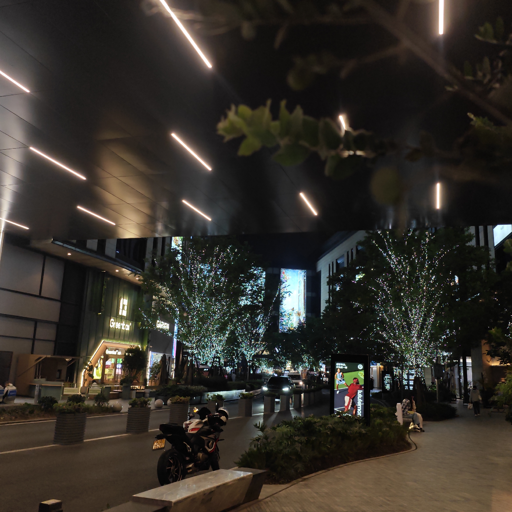 This image captures a vibrant night scene within an urban commercial district, likely in Yuzhong District, China. The setting features modern architecture illuminated by an array of artificial lights.
Above, a dark, angled canopy structure is adorned with multiple long, linear LED lights, creating a distinctive pattern and casting reflections. Beneath this overhead structure, a paved street or pathway leads through the area, flanked by buildings and lush greenery.
Numerous trees lining the thoroughfare are heavily decorated with countless small, sparkling string lights, creating a magical and festive atmosphere. Integrated into the building facades are large, brightly lit vertical display screens. One screen on the right prominently features an advertisement for "lululemon," showing two figures engaged in a sport, possibly tennis or badminton. Another building on the left displays a glowing green sign that reads "Greenbox," with "Greenbox Lab" also visible below it. A partial sign for "GARDEN" is also discernable.
In the foreground and middle ground, several subjects are visible. A black and white motorcycle is parked on the left side of the road, its license plate partially visible with "52Y0" showing. Further down the road, a dark-colored car with its headlights on is approaching. Potted plants and low shrubs are strategically placed along the walkways and road, adding to the urban landscape.
Several people are present in the scene, enjoying the evening. One person is sitting on a white bench near the "lululemon" display, while others can be seen walking or sitting in the background near the shops. The overall impression is that of a bustling and attractive evening destination, characterized by bright lights and modern design. The weather appears clear.