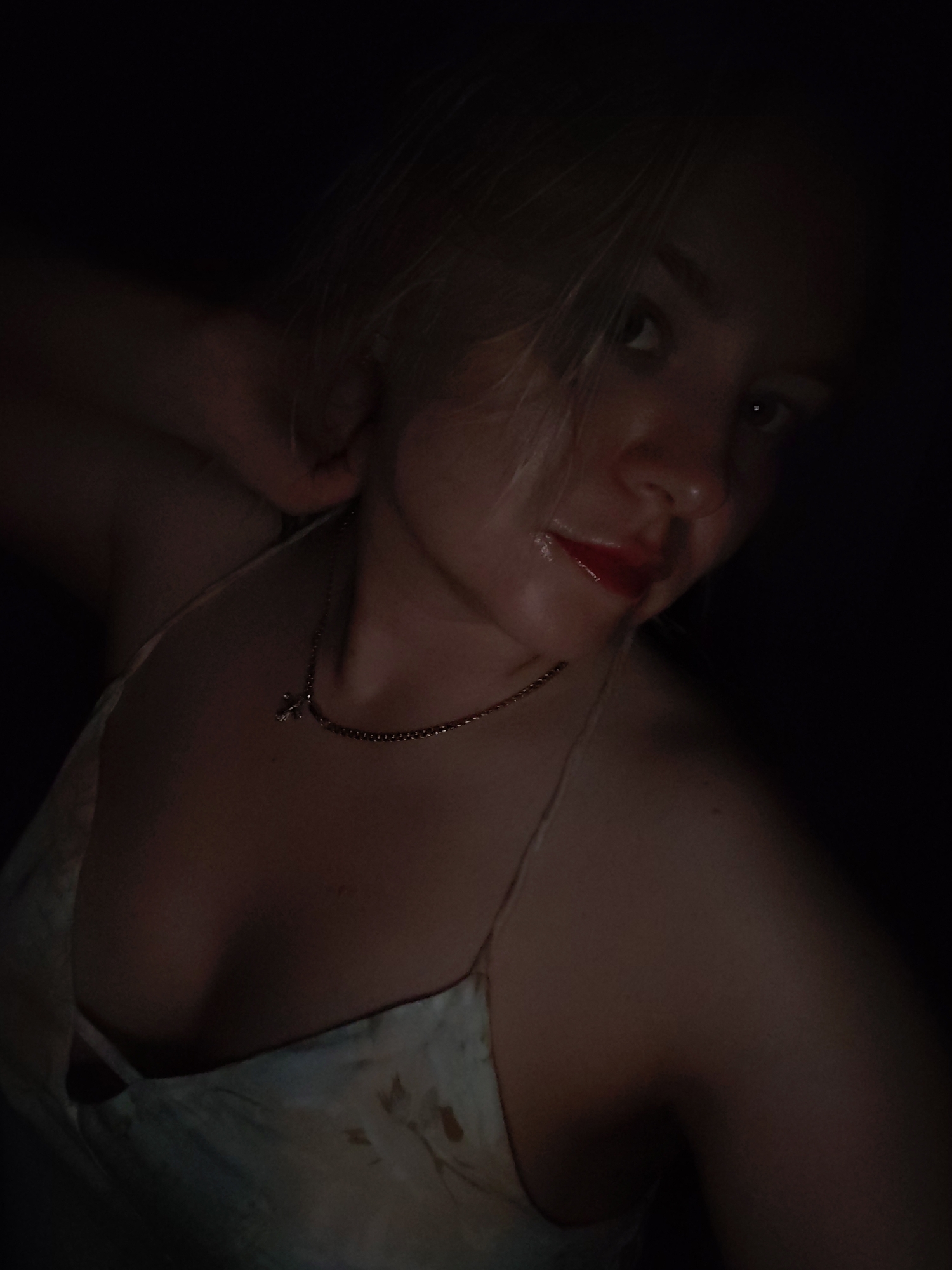 A dimly lit, close-up portrait of a young woman with fair skin and light blonde hair. She is wearing a light-colored tank top and a delicate gold necklace with a small cross pendant. Her lips are glossy with red lipstick. The background is dark and indistinct. The scene does not provide information regarding location.