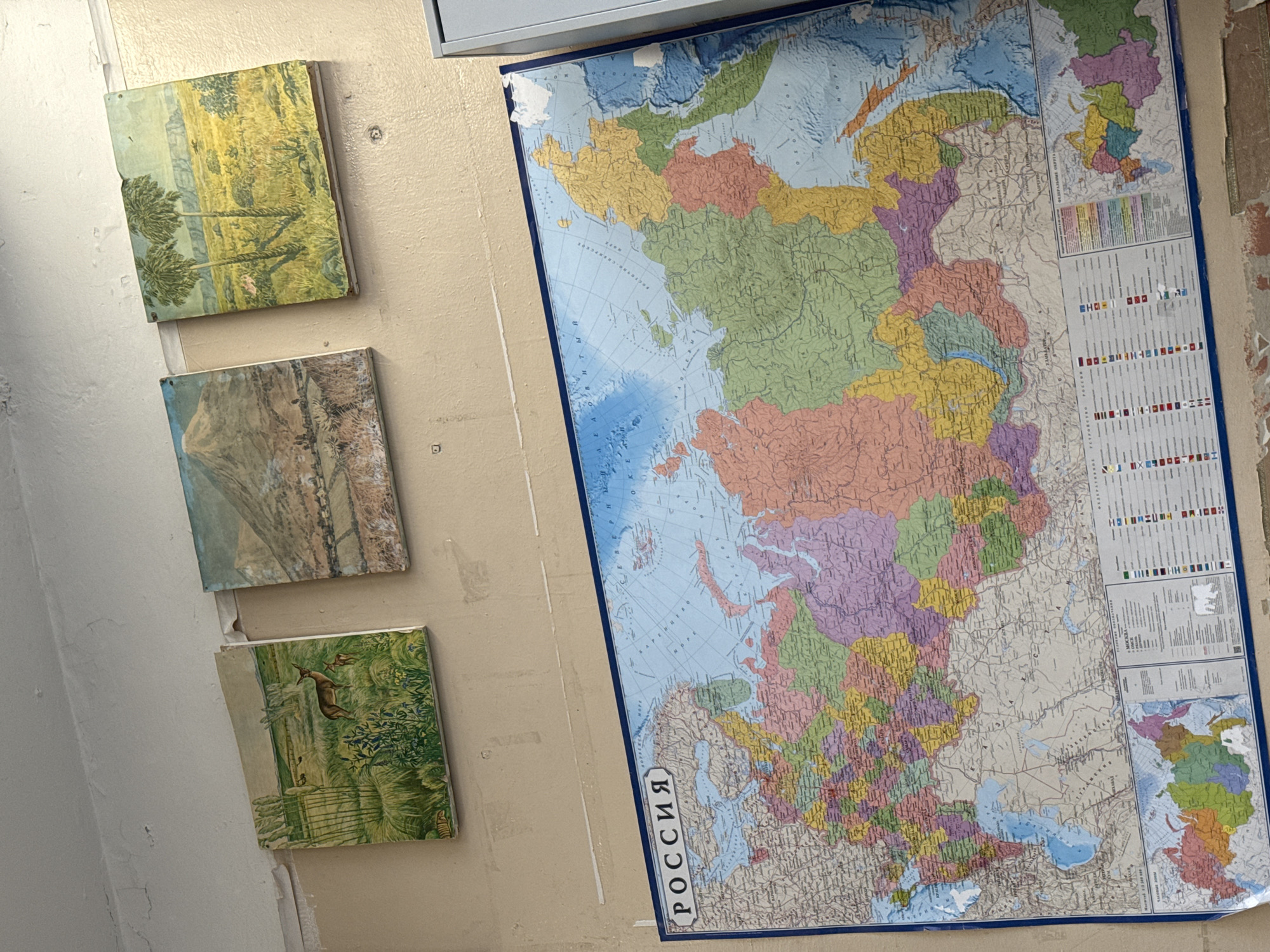 The image shows a section of a wall. Three square, framed pictures depicting outdoor scenes are arranged vertically along the left side. The top picture appears to show a field and trees. The middle picture contains a mountain. The bottom picture depicts a deer. To the right of the pictures, a large map of Russia hangs on the wall, its label visible. The wall is painted a light beige color. The scene does not suggest a specific location in Saint Petersburg.