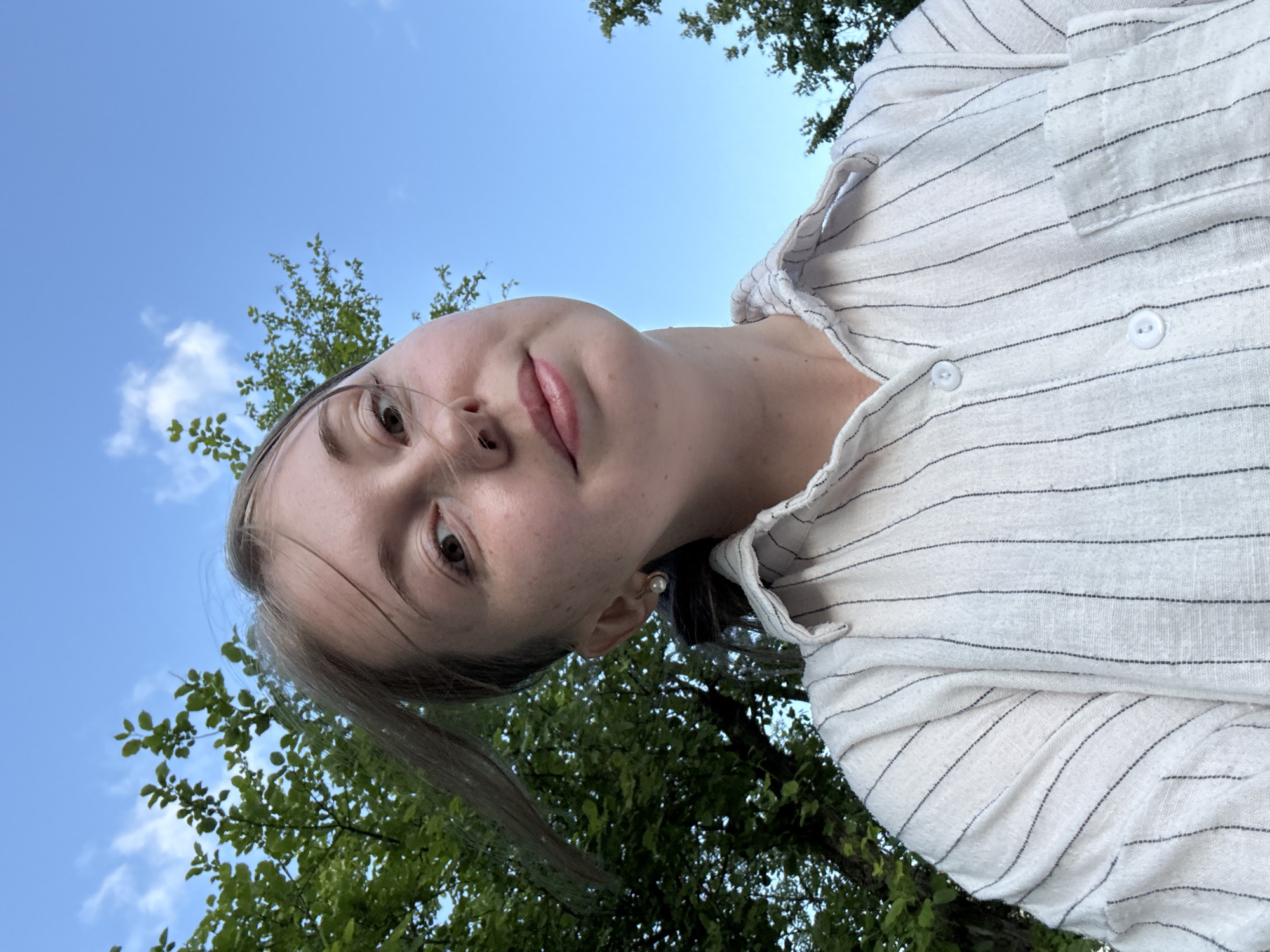 A medium shot shows a young woman with light brown hair and fair skin from a low angle outdoors. She is looking directly at the camera with a slight smile. Her hair is pulled back and some strands are blowing in the wind. She is wearing a white collared shirt with thin black vertical pinstripes and a pearl earring in her left ear. The background is a bright blue sky with some clouds, and the tops of green trees are visible on the left and right sides of the frame. The scene is outdoors, likely in a park or a backyard, under a clear sky. There is no indication of the location being in a city.