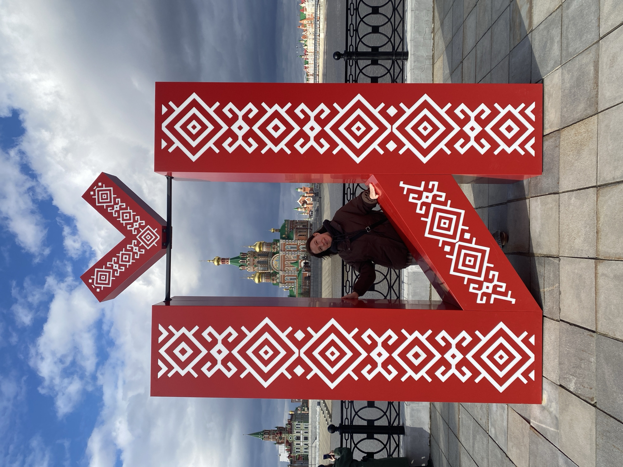 The image depicts a woman crouched within a large red Cyrillic letter "Ж" structure, adorned with white geometric patterns. The letter is situated on a paved area with a dark, wrought iron fence in the background. Beyond the fence is a body of water and various buildings, including a prominent Eastern Orthodox church with multiple golden domes. The sky transitions from blue with white clouds to a dark, stormy gray. The scene is likely in Yoshkar-Ola, Russia, due to the presence of the distinctive architectural styles. The woman wears a brown jacket and a black scarf.