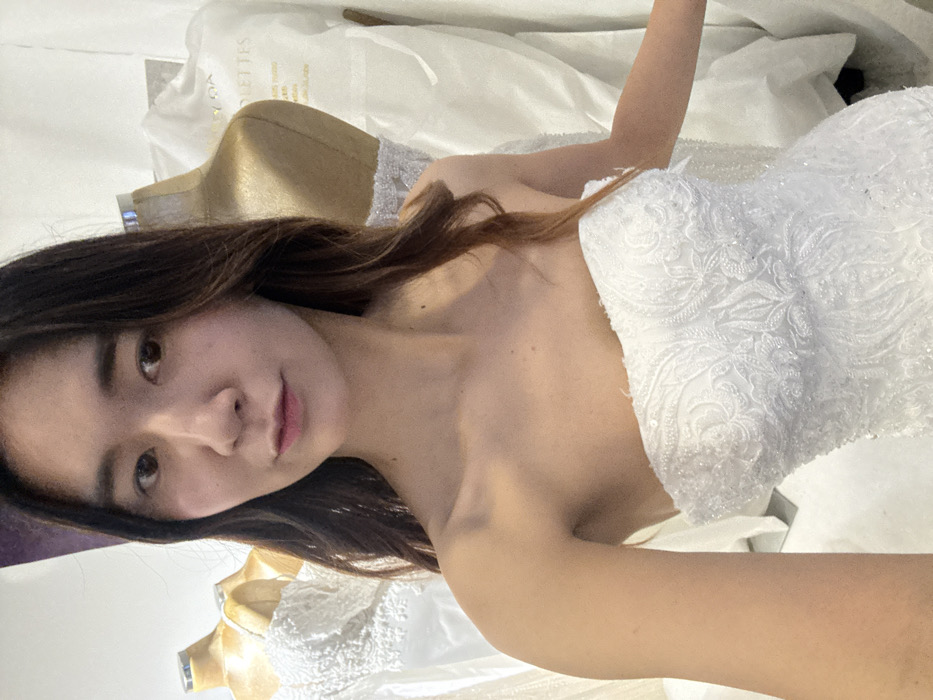 An adult female is depicted from the upper torso upwards, taking a selfie in what appears to be a bridal boutique. The individual has long, dark brown hair and is wearing a strapless white gown with intricate lace embroidery and beadwork. Her expression is neutral, and she looks directly into the camera. In the background, two headless mannequins are visible, each displaying a white dress. A white garment bag or cover hanging in the right background displays the gold text "VOLETTES" and "Mariage Sur-Mesure," identifying the location as "Volettes Paris," a bridal couture house located in Paris, France. The scene is brightly lit.
