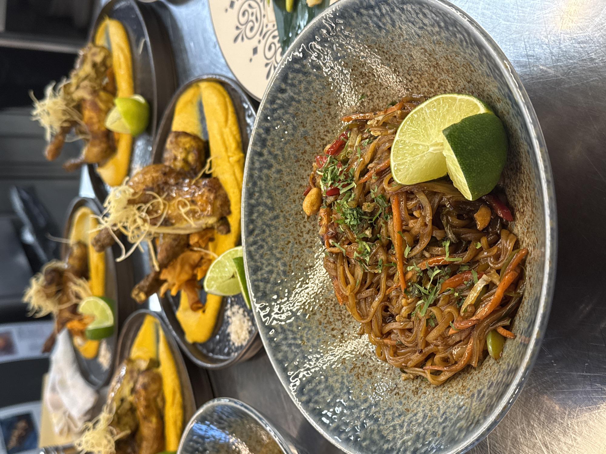 A close-up, high-angle shot shows a bowl of noodles on a stainless steel counter, likely in a restaurant kitchen. The noodles are thin and appear to be stir-fried with vegetables such as carrots and peppers. They are garnished with chopped herbs and topped with two lime wedges. In the blurred background, several plates of roasted chicken are visible, each served on a bed of orange puree and garnished with crispy threads and a lime wedge. The overall impression is of a gourmet meal, artfully presented. The exact location in Sofia, Bulgaria, is not discernible from the image alone.