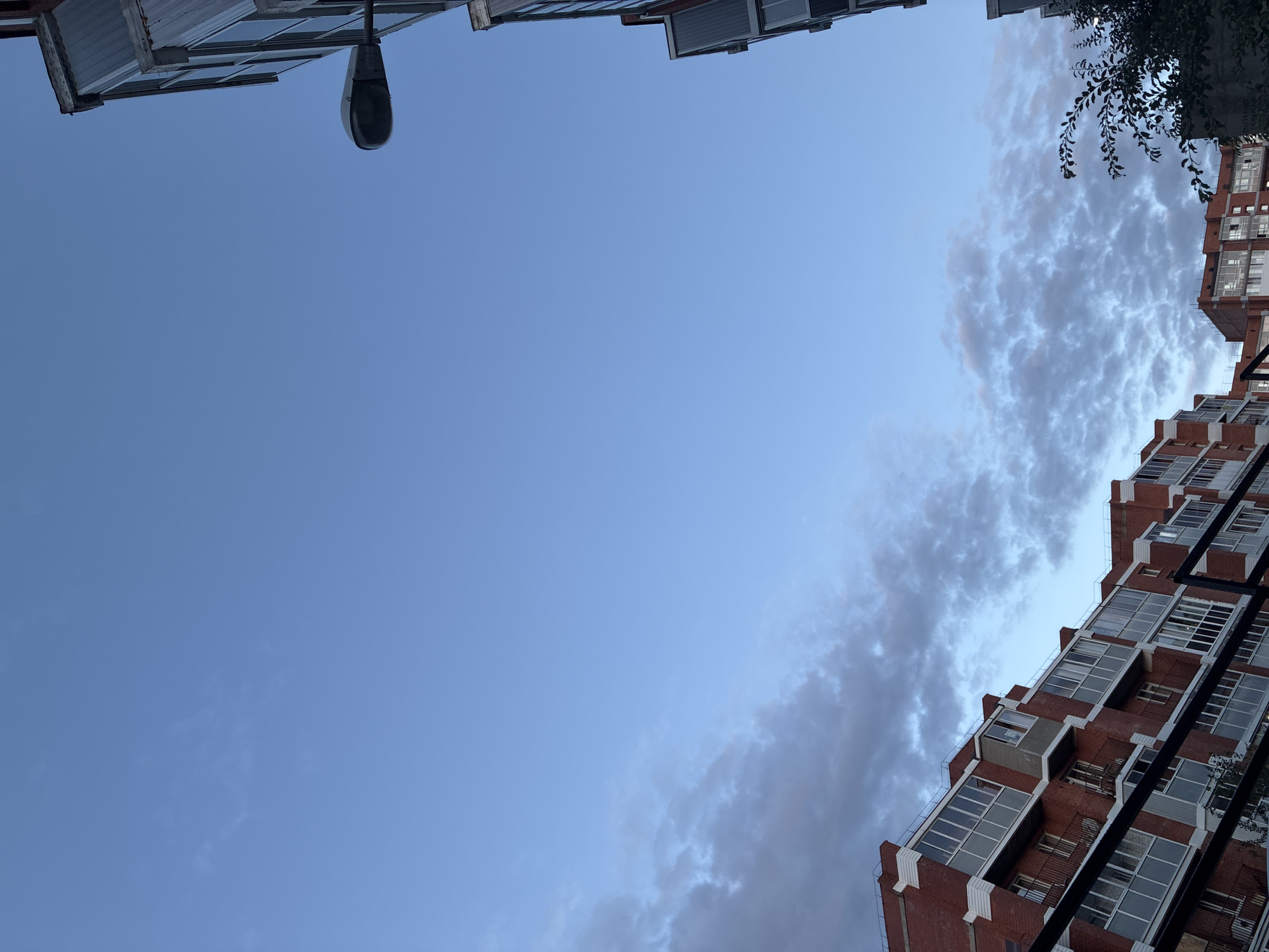 In the city of Markova, Russia, the image captures a clear blue sky with scattered, wispy clouds. The lower portion of the frame reveals the upper floors of multi-story brick buildings, characterized by numerous glass-enclosed balconies and windows. A street lamp is visible on the right side of the image, attached to the side of one of the buildings. A dark metal railing runs across the bottom of the frame, partially obscuring the view of the lower parts of the buildings and some foliage. The scene appears to be taken from a slightly elevated perspective, looking upwards.