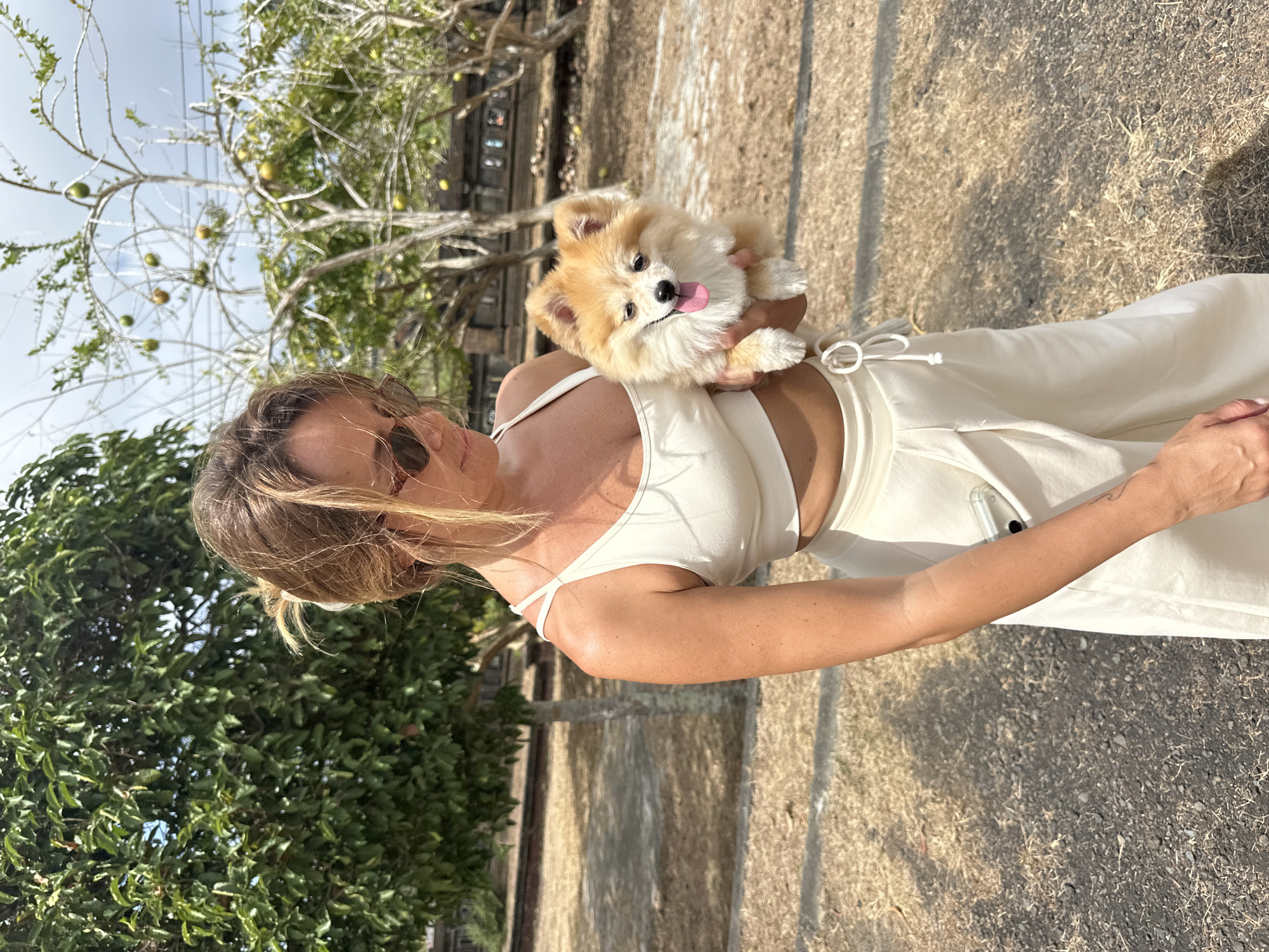 A woman with blonde hair pulled back and wearing sunglasses stands in an outdoor area, holding a small, fluffy, tan and white Pomeranian dog. She is dressed in a cream-colored crop top and matching loose-fitting pants. The dog is held in her left arm, facing forward with its tongue out. The background shows dry, patchy grass and dirt, with several trees. In the distance, there appears to be a stone structure with a railing and some greenery. The setting suggests a warm, sunny day in a park or historical site, potentially in Kuta Selatan, Indonesia, given the presence of tropical vegetation and architectural elements that could be found in such locations.