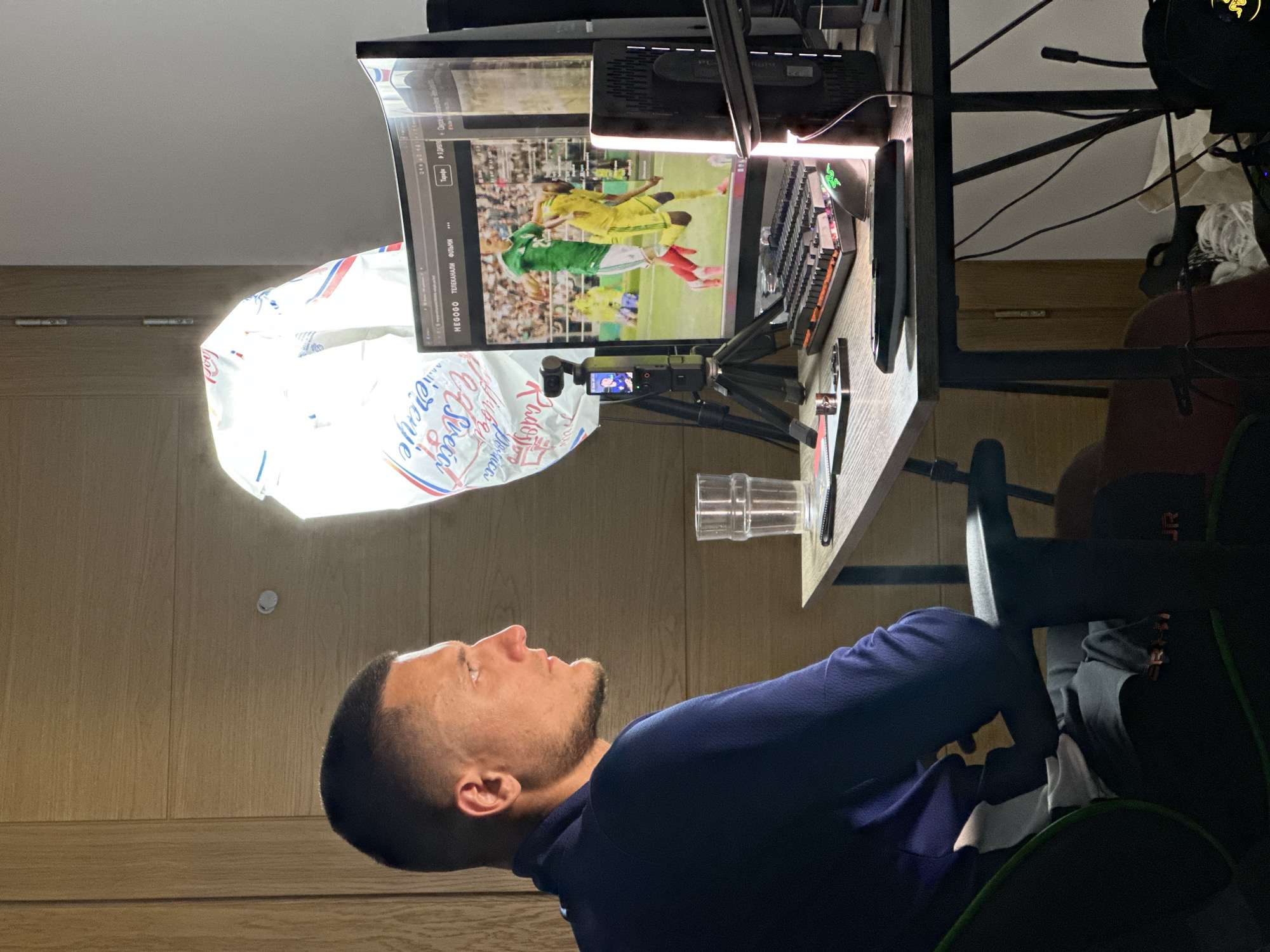 In a dimly lit room, a man with short, dark hair and a beard is seated in a chair, facing right. He is wearing a dark blue long-sleeved shirt. His gaze is directed towards a curved computer monitor displaying a soccer match. To the left of the monitor, a small camera on a tripod is positioned. On the desk, a glass of water, a smartphone, and a keyboard are visible. Behind the man and the desk, a large, illuminated softbox is present, suggesting it is being used for lighting. The background consists of a wooden door on the left and a light-colored wall on the right. The scene appears to be set in a private residence, possibly in Donji Pobori, Montenegro, given the provided context.