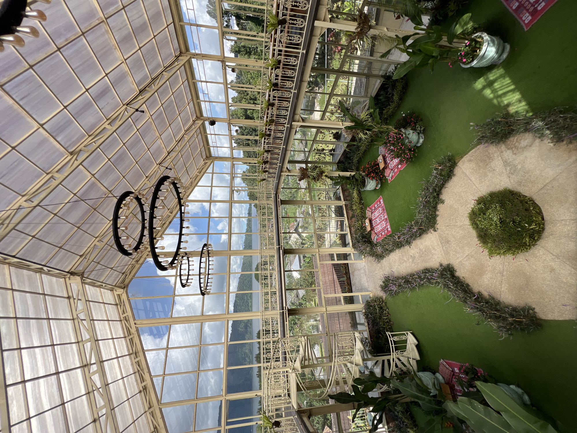 The image shows the interior of a building with a glass roof, likely a greenhouse, located in Baturiti, Indonesia. The roof is supported by a framework of light-colored metal beams. Circular chandeliers with multiple lights hang from the ceiling. The windows display an outdoor view of mountains, trees, and a lake. The interior space features a green lawn, a beige stone walkway, and a circular garden with a shrub in the center. Small potted plants and decorative items are arranged around the lawn area. A balcony with white metal railings runs along the interior walls, partially obscured by plants. Two red carpets with inscriptions are placed along the pathway. White metal chairs are situated near the plants.