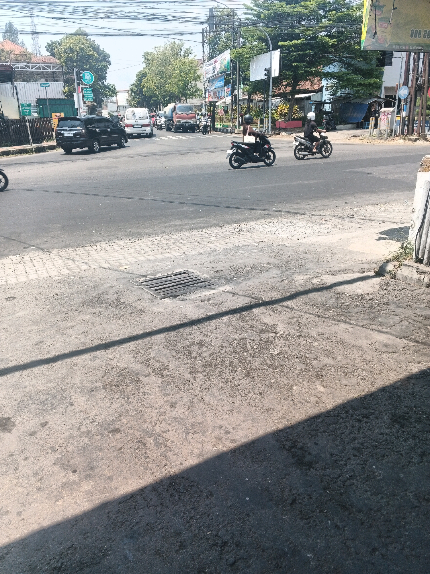 This is a street-level, eye-level, full-shot, outdoor, daytime photograph taken at an intersection in Ungaran, Indonesia. The scene depicts typical urban traffic, including cars and motorcycles. In the foreground, a portion of the road and sidewalk is visible, with a drain grate present on the sidewalk. The middle ground of the image shows a busy intersection with various vehicles, including a black car in the left lane, a white van and a red truck in the right lanes. Two individuals on motorcycles are also visible, one in the left lane and another in the right lane, both appearing to be moving through the intersection. In the background, buildings and lush green trees line the street, and power lines are strung overhead, indicating an urban environment. The lighting suggests a bright, sunny day.