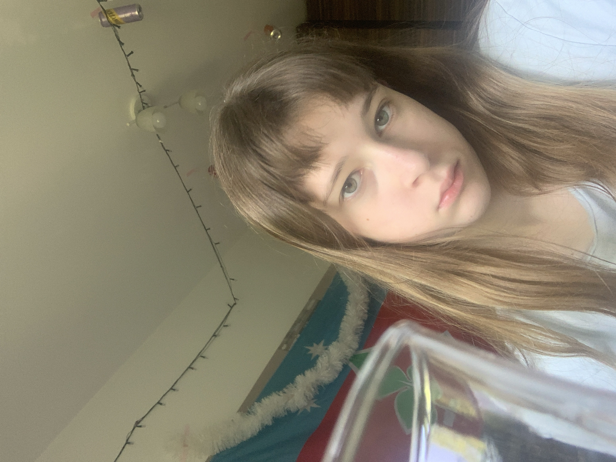 A young woman with light brown hair and bangs is looking at the camera. She is wearing a light-colored shirt. The background includes a wall with string lights and a decorative flag with white garland. A can is attached to the string lights. Part of a piece of furniture is visible in the upper right corner. A glass with green is in the lower right part of the image. Location is indeterminate, and there is no definitive indication of the city of Vozdovac Urban Municipality, Serbia.