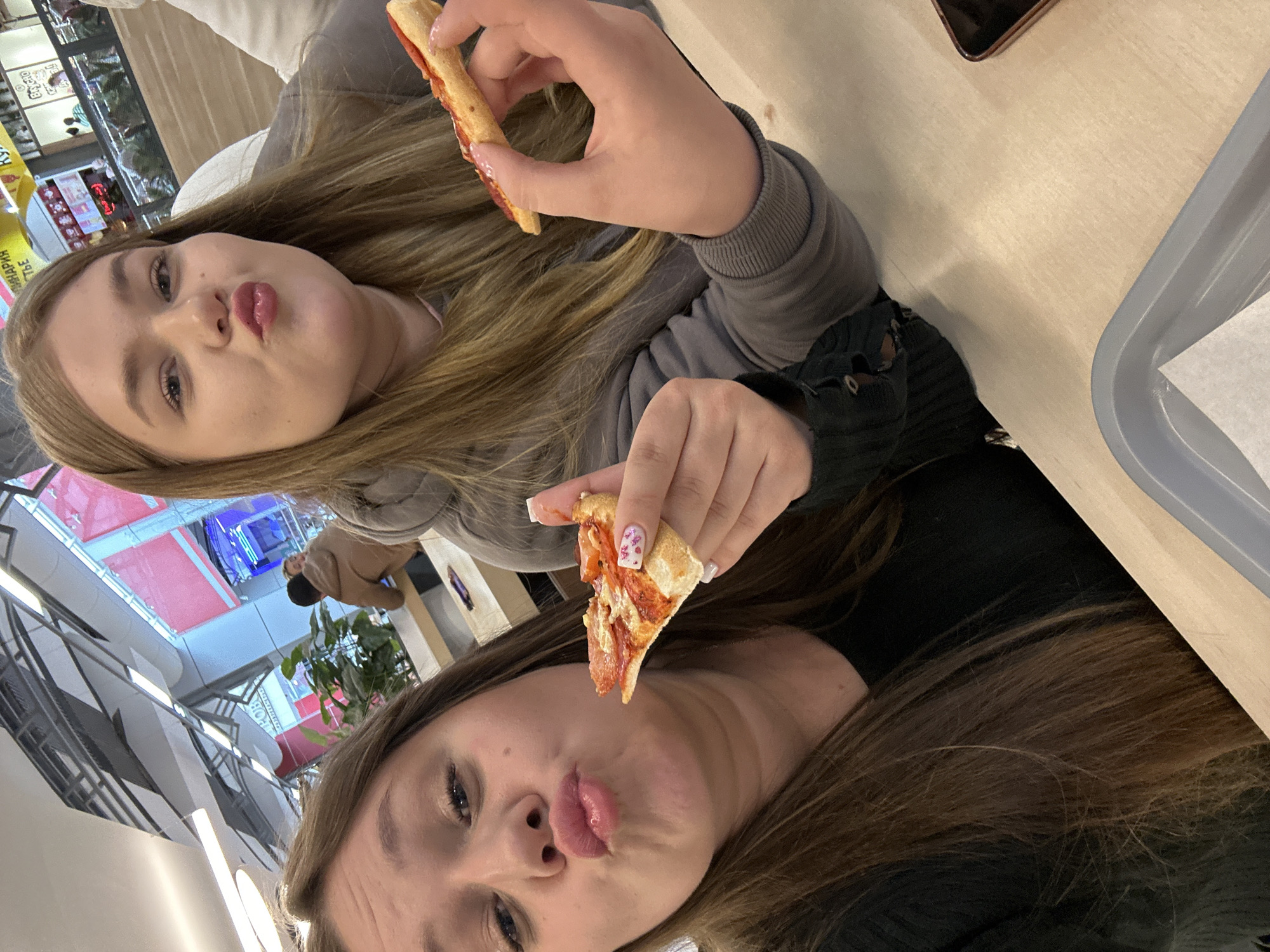 Two young women are posing for a selfie while holding slices of pizza. The woman on top is wearing a gray sweatshirt, and the woman on the bottom is wearing a black top. They both have blonde or brown hair and are making a duck-face expression. The image seems to be taken in a food court setting, with a tray and a table visible. In the background, there is a view of the interior of a shopping mall or similar establishment, identifiable by elements typical to food courts and retail spaces. There is no visible detail that can identify the location in the city of Izhevsk, Russia.
