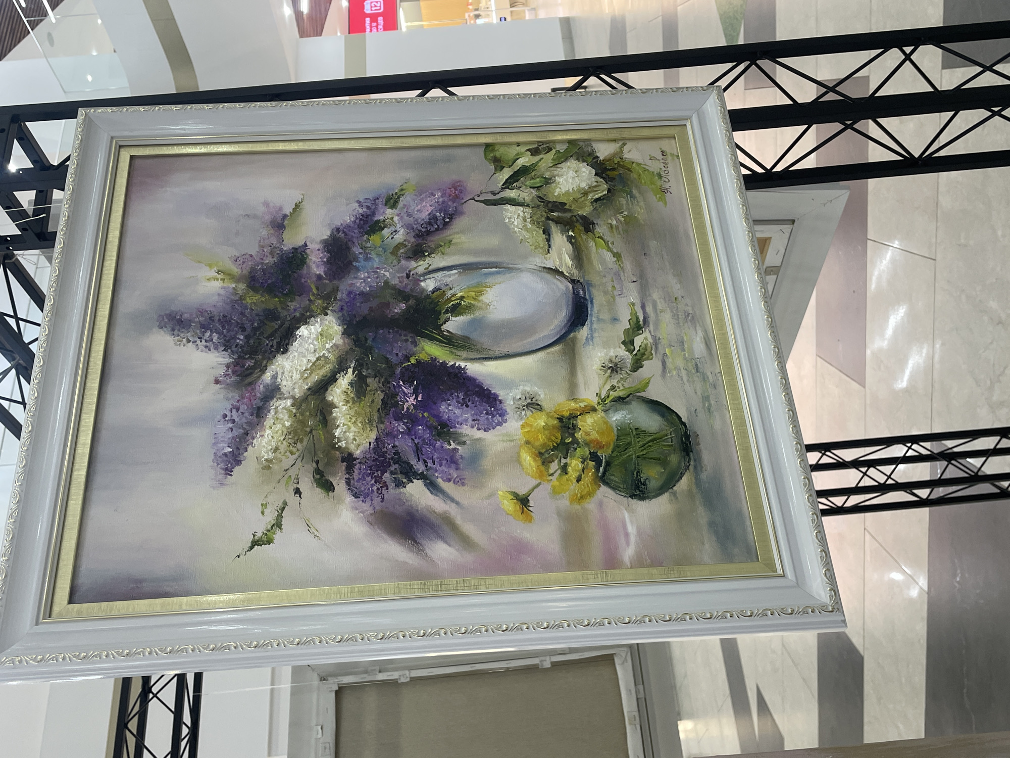 The image shows a painting of a floral still life. The painting features a bouquet of lilacs in shades of purple and white arranged in a clear glass vase. To the right of the vase, there is a smaller, green glass vase containing a cluster of yellow flowers. The background is a soft, blended gradient of light colors, creating a gentle atmosphere. The painting is framed in an ornate white and gold frame and is displayed on a black metal stand. In the background, there is a glimpse of a mall interior, indicated by polished floors, retail displays, and architectural elements, suggesting the painting is part of an exhibition or sale within a commercial space in Kokshetau, Kazakhstan. No people are clearly visible in the image.