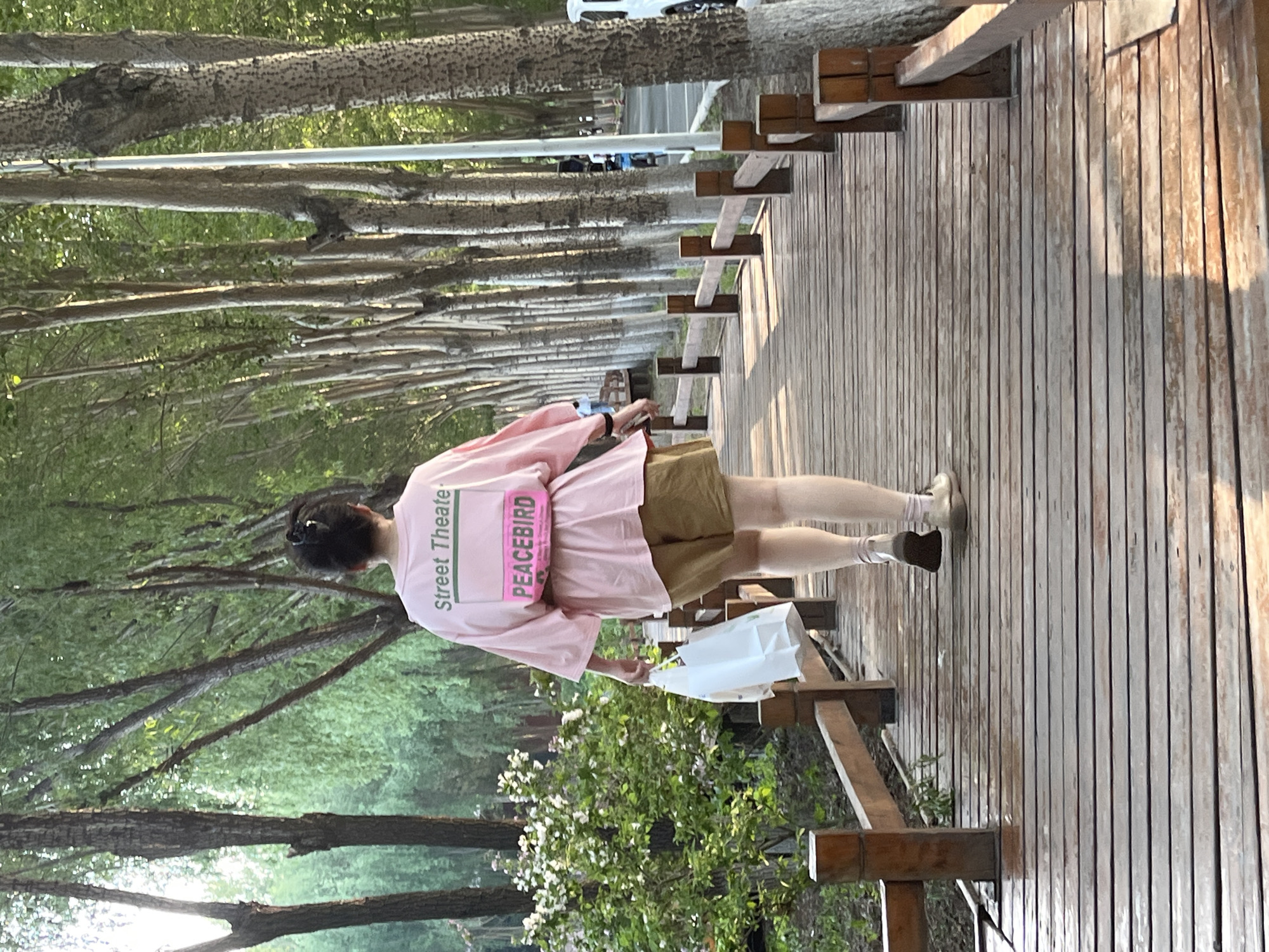 A medium shot photograph of a woman standing on a wooden bridge on an alleyway in Changchun City, China. She has brown hair pulled into a bun and is wearing a pink t-shirt with “Street Theatre” printed vertically on the upper left back and “PEACEBIRD” in a pink rectangle in the middle back of her shirt. She is wearing khaki shorts and a beige sock and shoe combination. She is holding a white shopping bag in her left hand. The wooden bridge she is standing on is comprised of thin planks of woods, and is raised off the ground. There is a wooden railing on the right-hand side of the bridge, and the bridge is surrounded by overgrown bushes and trees. The alleyway in the background is lined with tall trees.