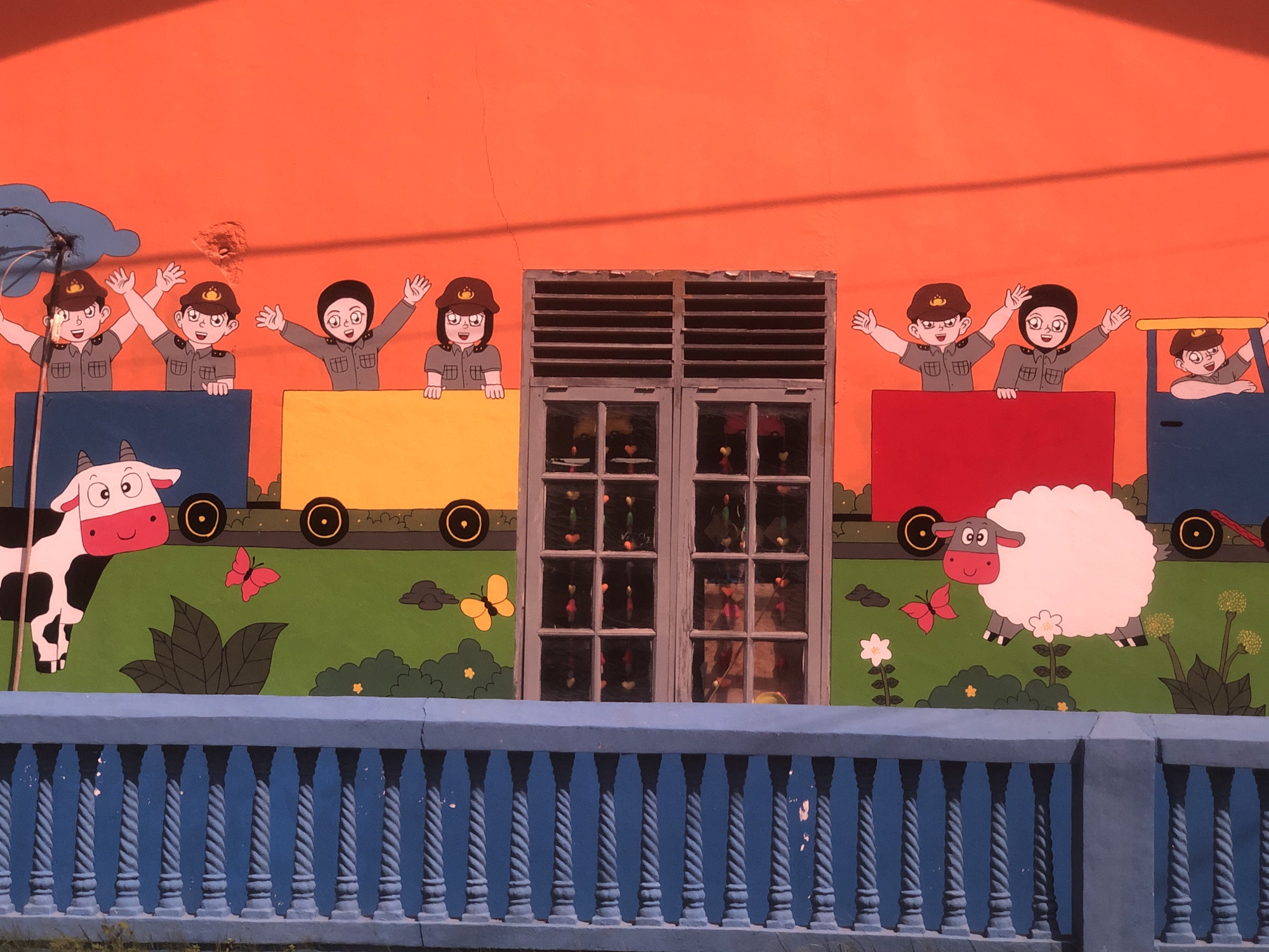 The image depicts a painted mural on a building facade in Lhokseumawe, Indonesia. The mural features a cartoon train comprised of a blue locomotive and three colorful cars (yellow, red, and blue). Cartoon children wearing hats are depicted riding in the train cars, waving. Below the train, painted on a green field, are a cow, a sheep, and butterflies. The building has a window in the center of the mural. A blue railing runs along the bottom of the image, in front of the mural.