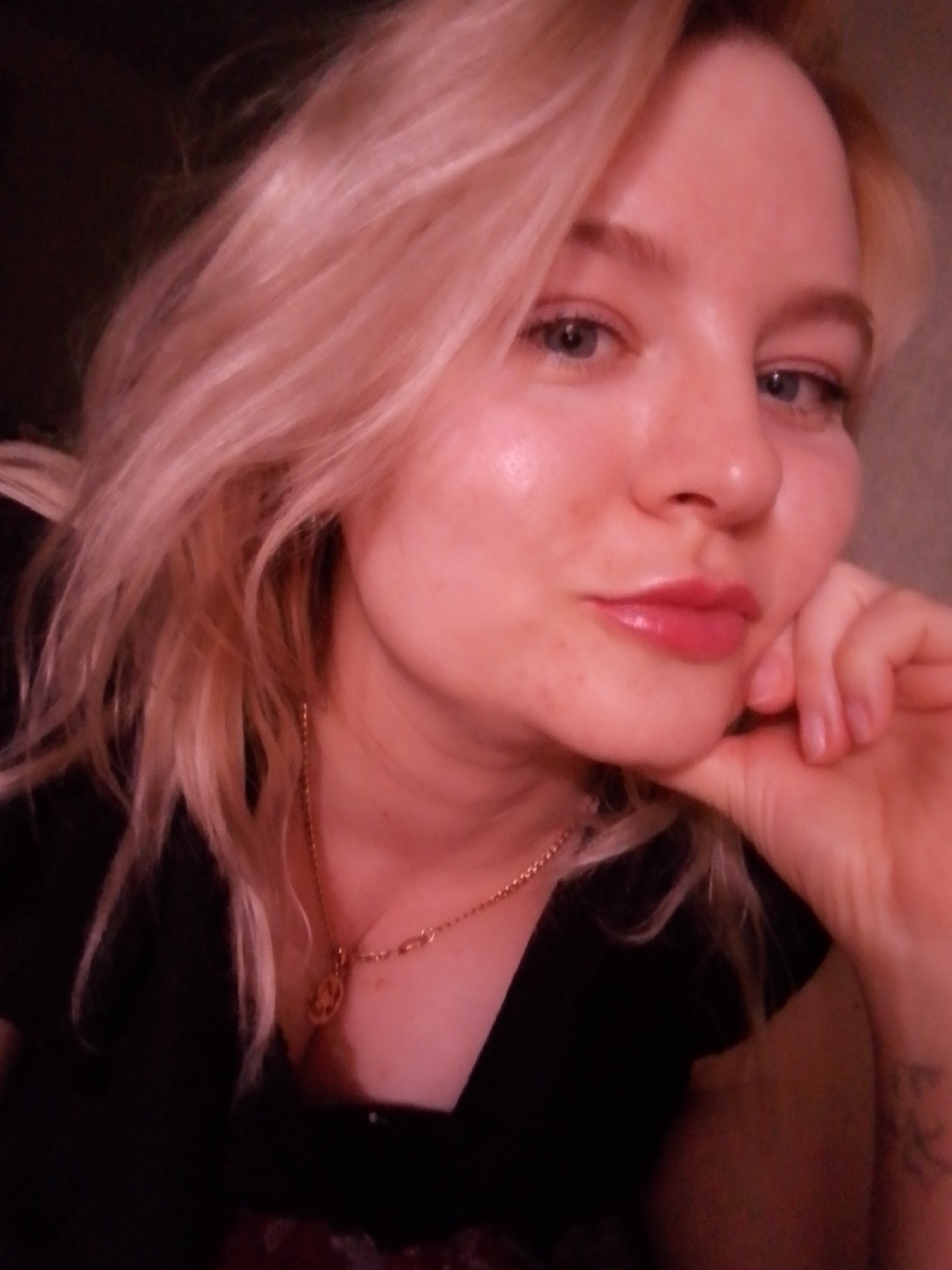 The image displays a close-up portrait of a female individual with fair skin, light blonde hair, and blue eyes. She is positioned with her chin resting on her right hand, which is visible up to the wrist. Her lips are a glossy pinkish-red. The individual wears a dark-colored garment with a low neckline and a gold-toned chain necklace with a circular pendant. Minor skin blemishes are visible on her chin and right cheek. A faint, indistinct tattoo is partially visible on her left forearm. The background is uniformly dark and out of focus, suggesting an indoor setting with low ambient lighting. No visual cues within the image provide information about a specific location within the city of Izhevsk, Russia.