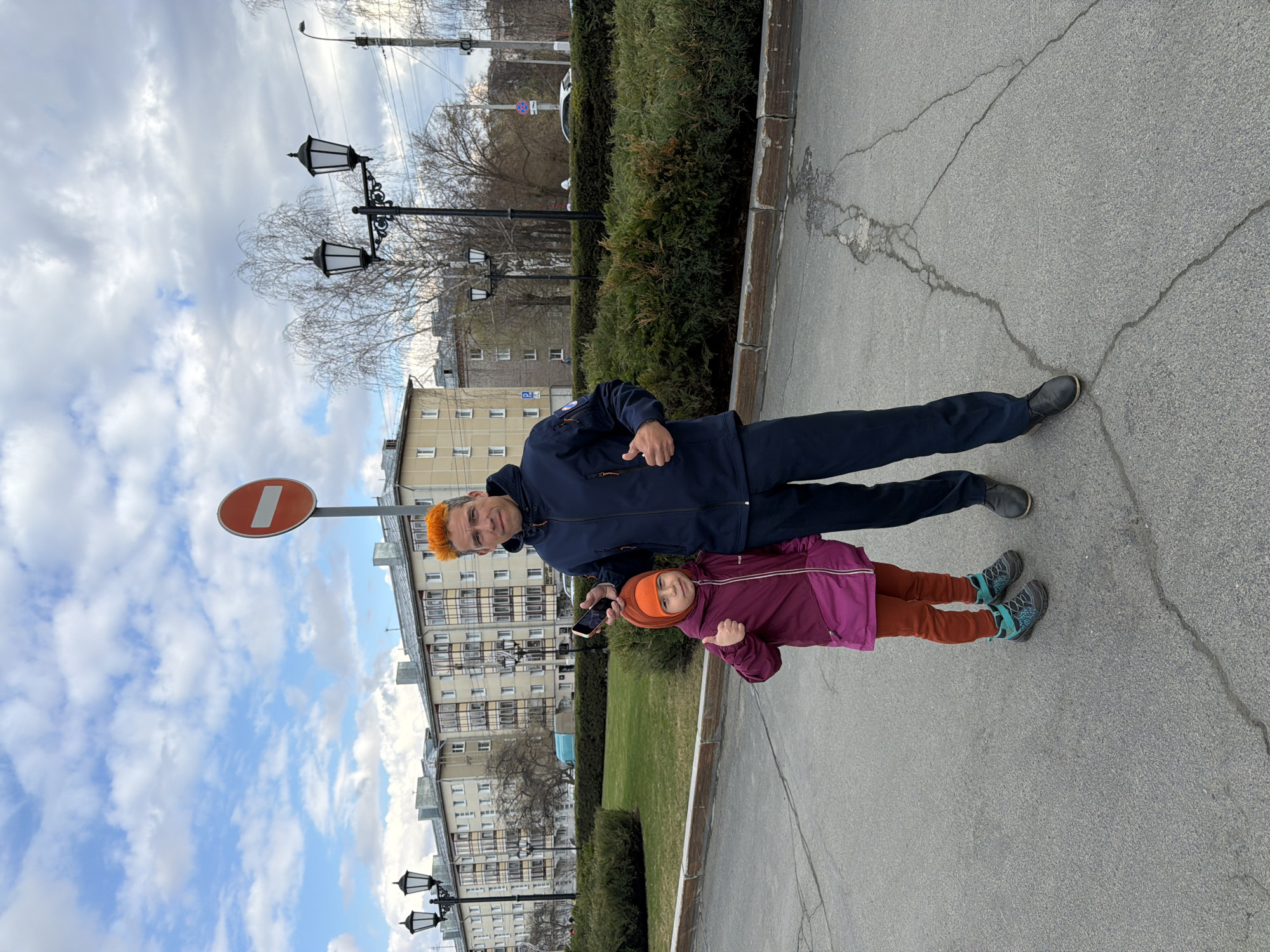 The photograph shows an adult male and a child standing on a sidewalk in Izhevsk, Russia. The man has orange hair and is wearing a dark blue jacket and dark blue pants. The child is wearing a burgundy jacket and orange pants. A "Do Not Enter" sign is visible in the background. The background also features buildings, trees, and ornamental street lamps, typical of urban settings. The sky is partly cloudy. The man is holding a mobile phone, possibly taking the picture.