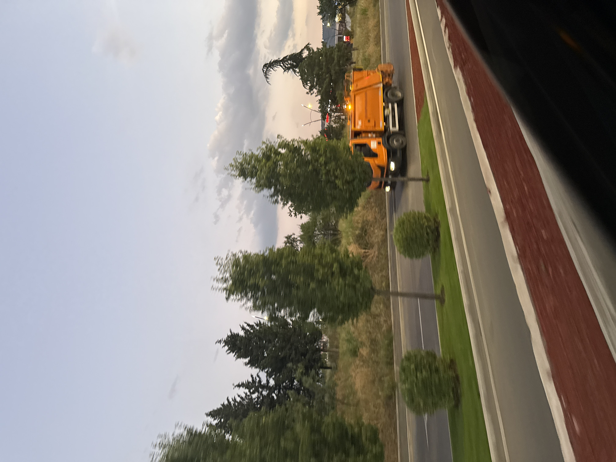 An orange garbage truck is seen driving on a road in Tbilisi, Georgia. The truck is moving from left to right in the frame. The sky is cloudy and appears to be dusk or dawn. There are several trees and some dry grass lining the road. In the foreground, there is a dark object which could be the interior of a vehicle, suggesting the photo was taken from a moving car. The overall scene is urban, with hints of industrial or commercial structures in the distance.