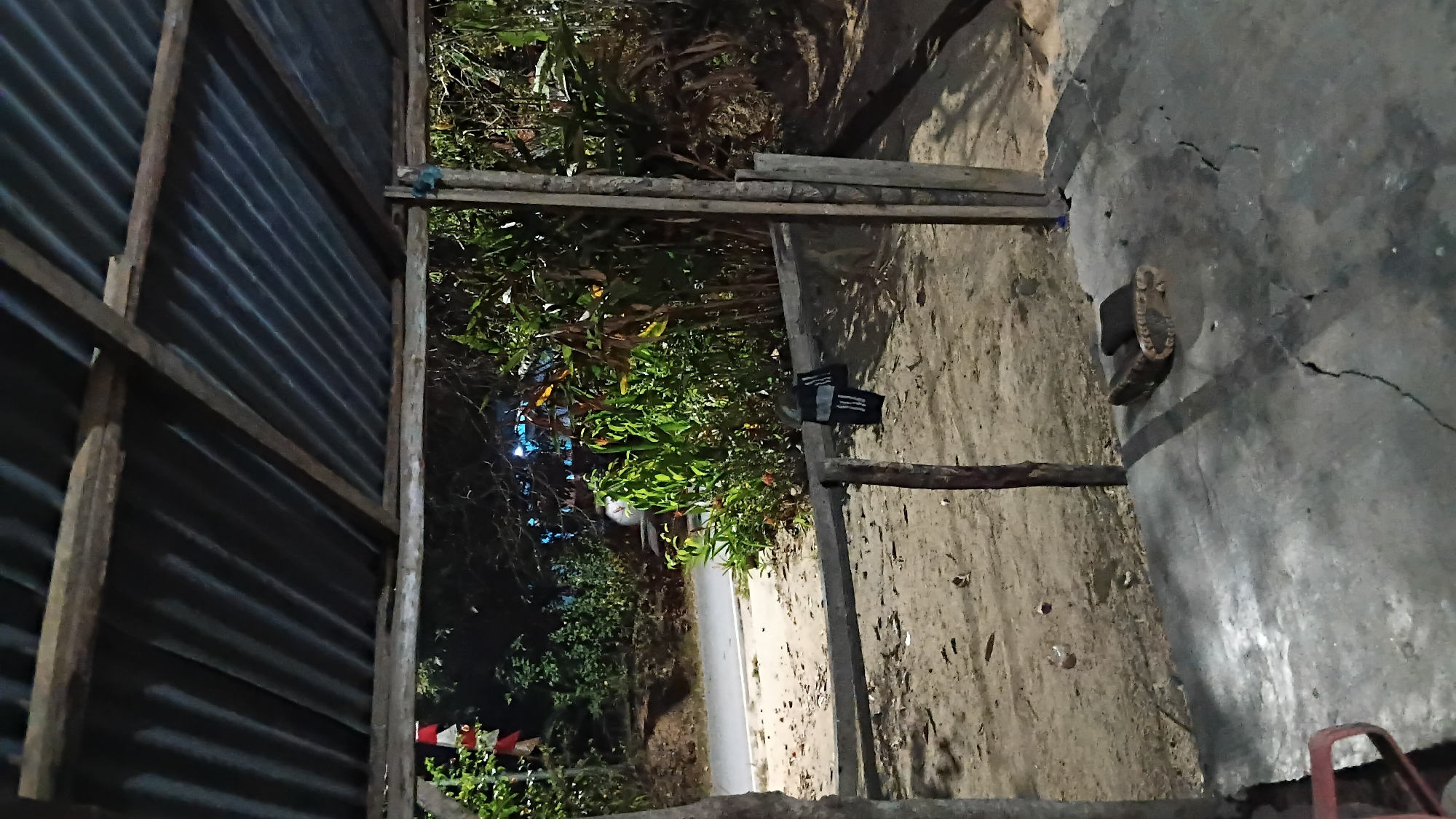 The image depicts an outdoor shelter with a corrugated metal roof supported by wooden beams. The floor is concrete, showing cracks. In the foreground, on the concrete floor, is a dark-colored, rectangular object, possibly a block or a shoe. A red chair is partially visible on the far left. The shelter opens to an area with sandy ground and sparse vegetation. In the midground, a dark-colored garment, appearing to be trousers or shorts with white stripes, is hanging from a wooden beam. Beyond the shelter, a road is visible with some greenery and trees. The scene appears to be set at dusk or at night, with artificial lights in the background. The overall impression is of a rustic, utilitarian structure in a semi-rural or suburban setting.