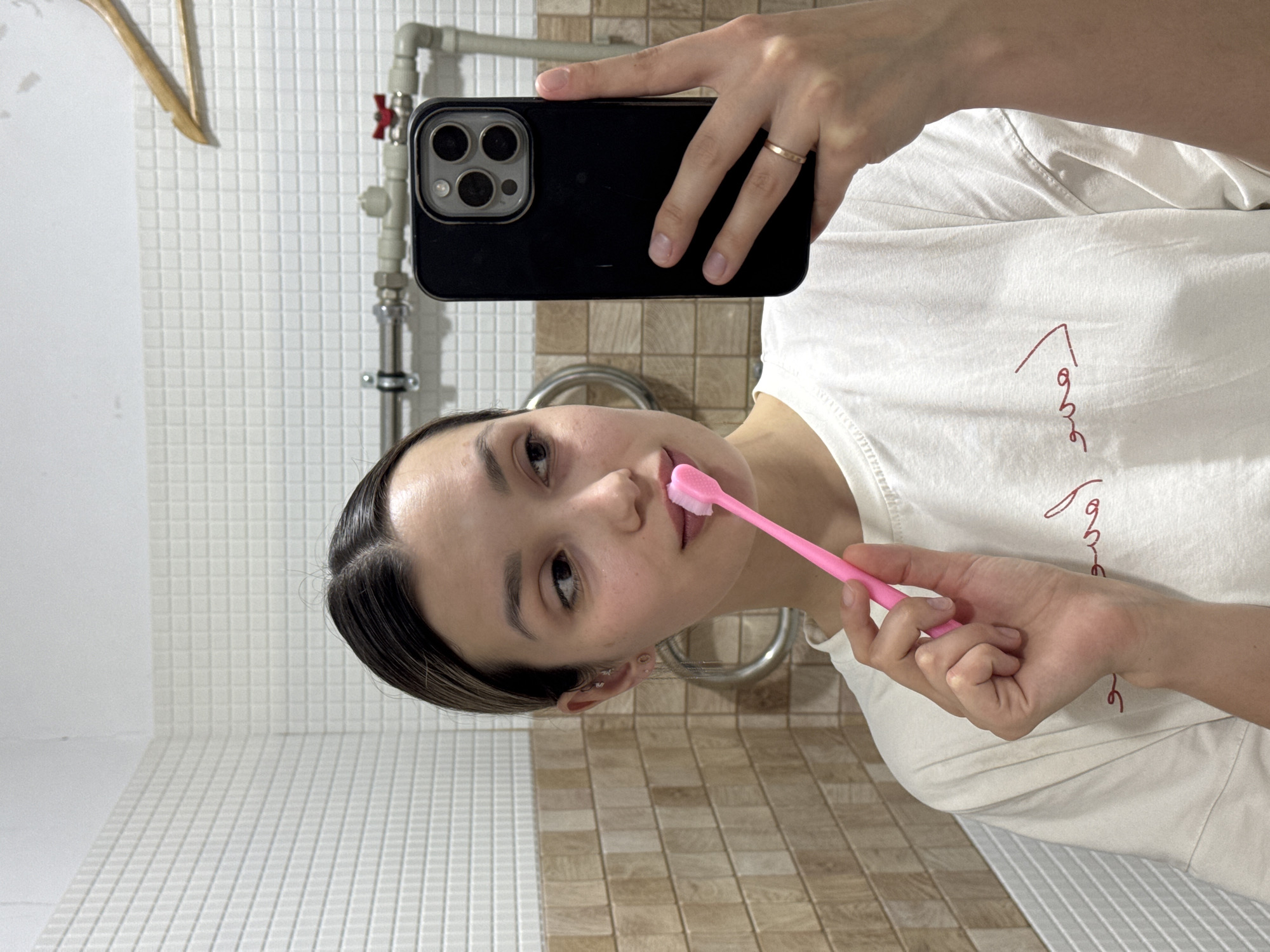 A young woman is depicted taking a mirror selfie in what appears to be a bathroom. The image is rotated 90 degrees counter-clockwise, making her appear sideways. She has dark hair, slicked back, and a neutral expression. She is wearing a white t-shirt with some red cursive writing, which includes an upward arrow design, visible on the front.
In her right hand, she holds a black iPhone, identifiable as a Pro Max model by its camera array, which she uses to capture the selfie. On her right ring finger, she wears a simple, thin gold-colored ring. With her left hand, she holds a pink toothbrush to her mouth, suggesting she is engaged in or about to begin brushing her teeth.
The background features two types of tiled walls: the upper portion consists of small white square tiles, and the lower portion has larger, beige and brown rectangular tiles. Visible behind her are exposed pipes with a red valve, indicative of plumbing, and a chrome-plated towel rack. The lighting is consistent with an indoor setting.