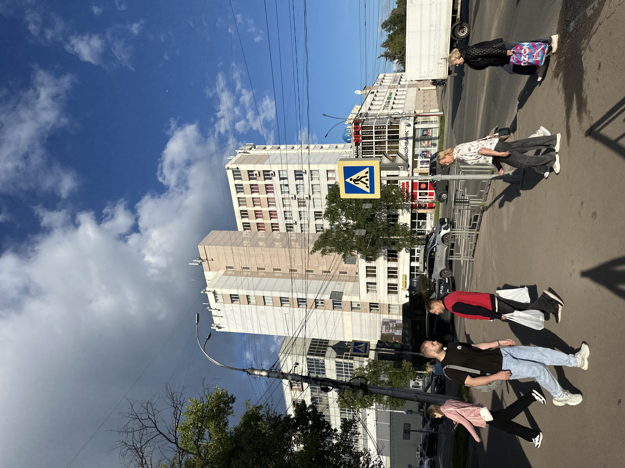 In Rubtsovo, Russia, on a bright day with a mix of clouds and blue sky, a wide street is bustling with activity. Pedestrians cross a street at a marked crosswalk in front of a large, modern multi-story building with a light-colored facade. Several people are walking, including a woman in a pink jacket, a man in a black t-shirt and light blue jeans carrying a backpack, and another man in a red and black jacket carrying white shopping bags. Two women are walking away from the viewer, one in a patterned shirt and dark pants, and the other in a black jacket carrying a colorful shopping bag. Cars and a white truck are visible on the street. The environment is urban, with power lines crisscrossing the sky and trees lining the sidewalk.