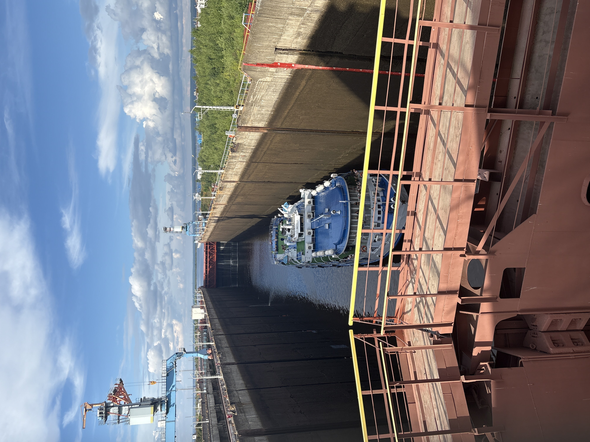 The image shows a large cargo ship entering a lock chamber on a sunny day. The lock walls are made of concrete and are very high. A blue and white passenger ship is already inside the lock, and it appears to be moving. There are cranes and other industrial structures visible in the background, as well as lush green trees. The location is the Votkinsk Hydroelectric Power Station lock, part of the Kama River water system, near the city of Chaikovsky, Russia.
