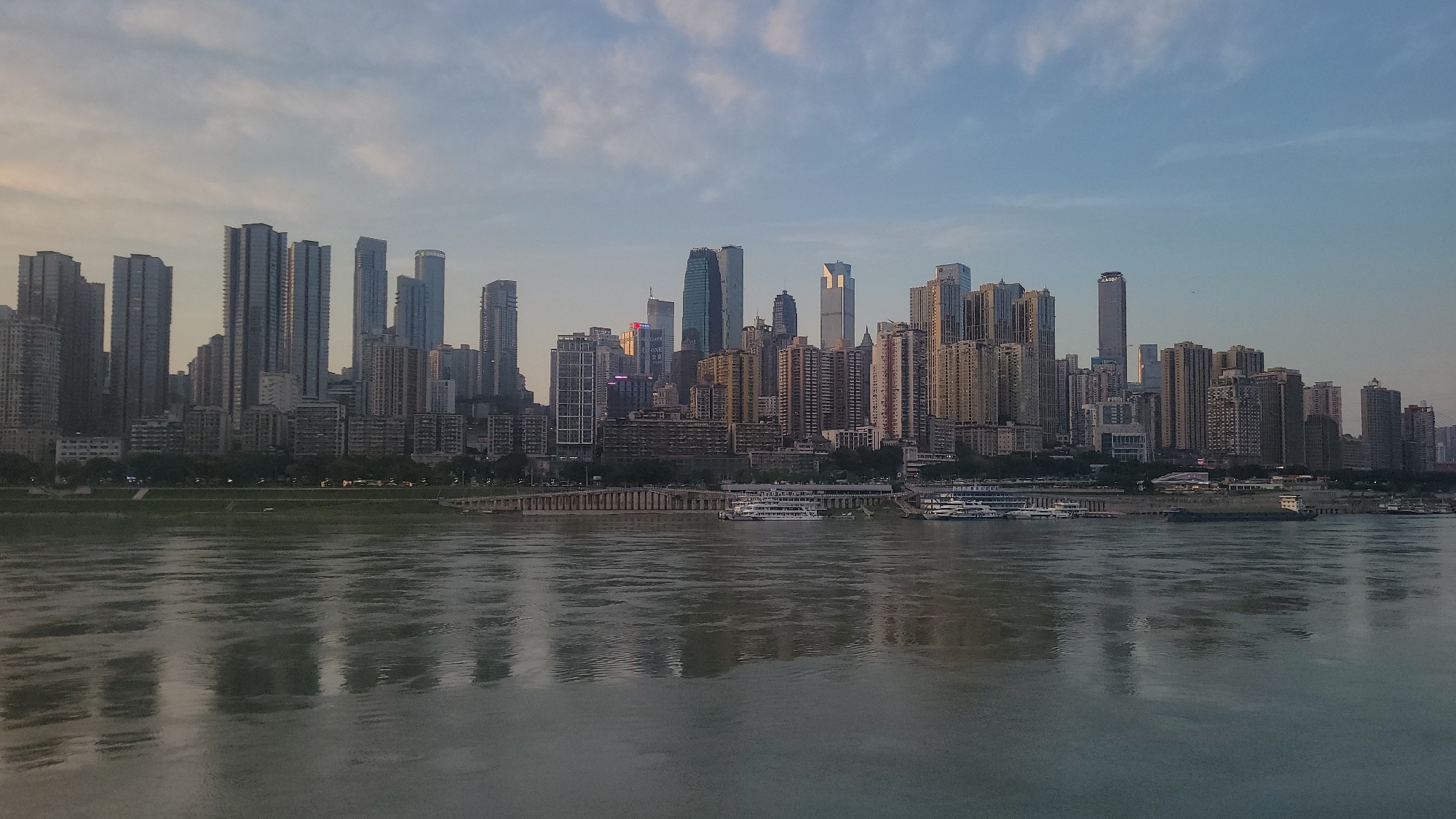 The image depicts a cityscape, likely taken from across a body of water. The skyline features numerous high-rise buildings of varying heights and architectural styles. The buildings appear to be densely packed together. In the foreground, the water's surface is calm with light reflecting off it. The sky is visible above the buildings, and the lighting suggests it may be either early morning or late evening. Several boats are docked along the shoreline.