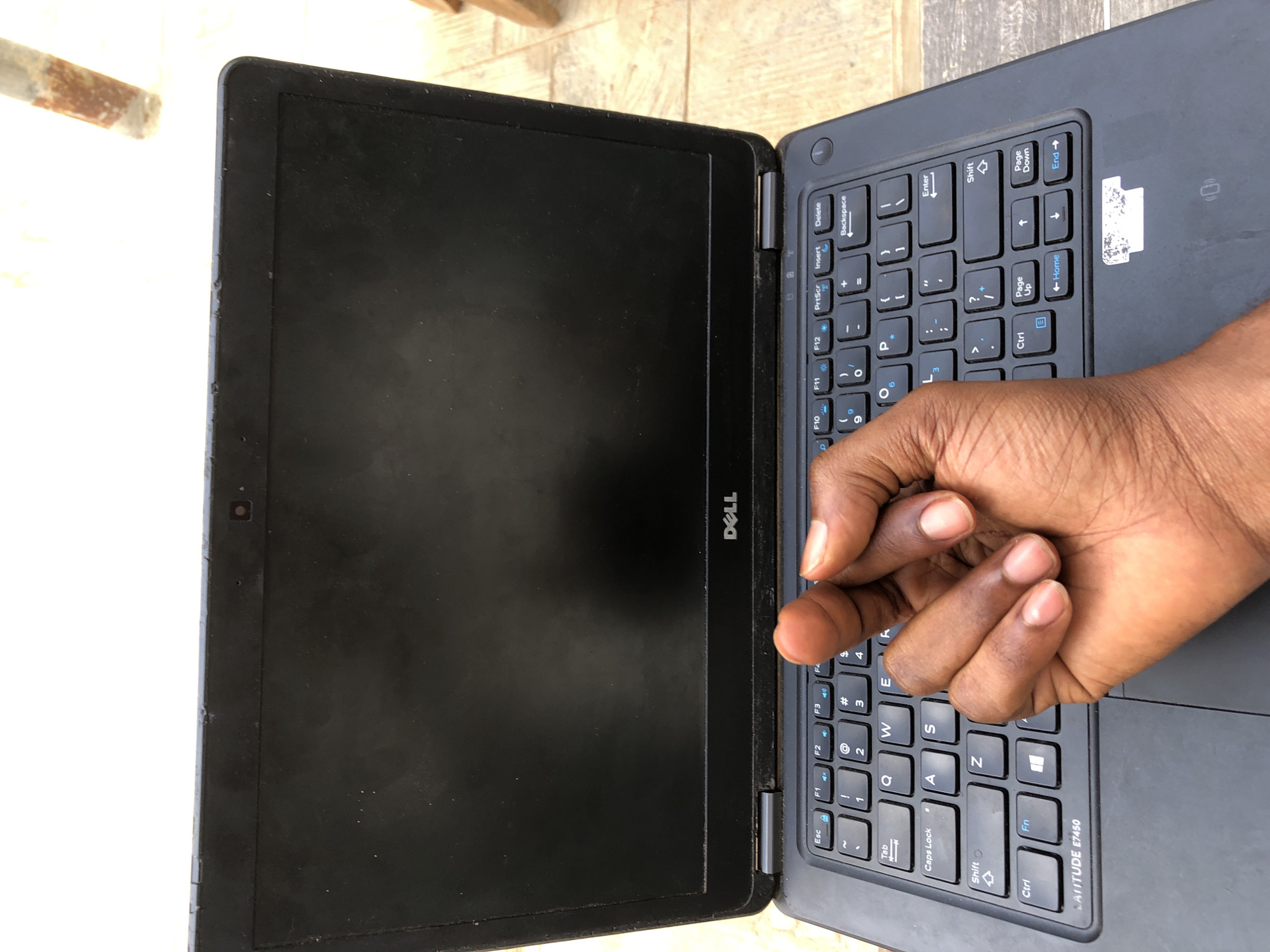 A partially open Dell Latitude E7450 laptop sits on a surface with a light-colored pattern, possibly a tile or wooden floor. The laptop's screen is black. A dark-skinned hand is resting on the keyboard. The keyboard layout is visible. A sticker is affixed to the laptop next to the arrow keys.
